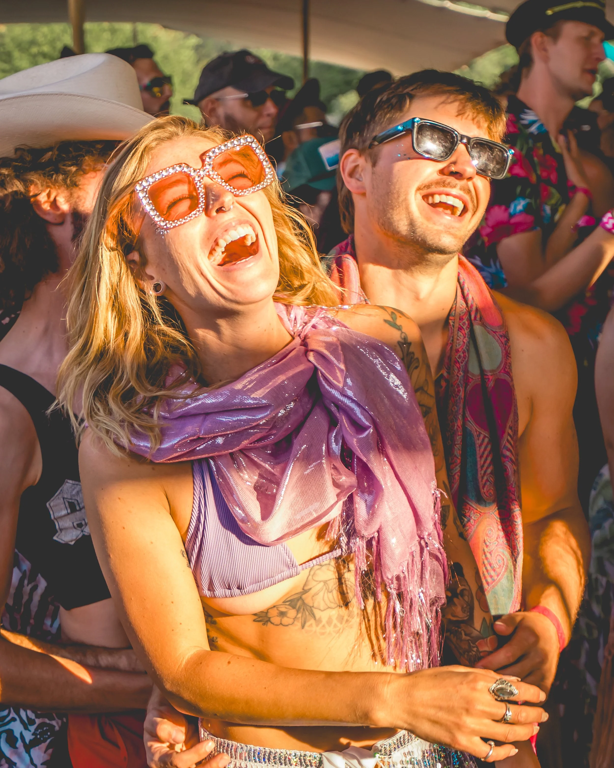 A group of people enjoying a lively outdoor event or festival, with a woman wearing oversized rhinestone sunglasses, a tied-up top, and a shiny pink scarf, laughing with a man in sunglasses and a sleeveless shirt.