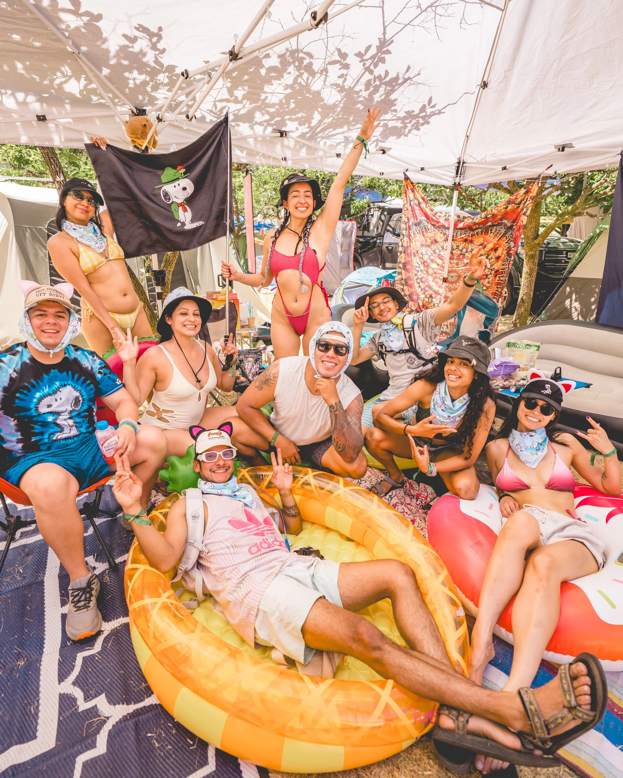 Group of friends at a summer camping trip, posing under a canopy with outdoor tents, decorations, and inflatable pool floats, celebrating and smiling.