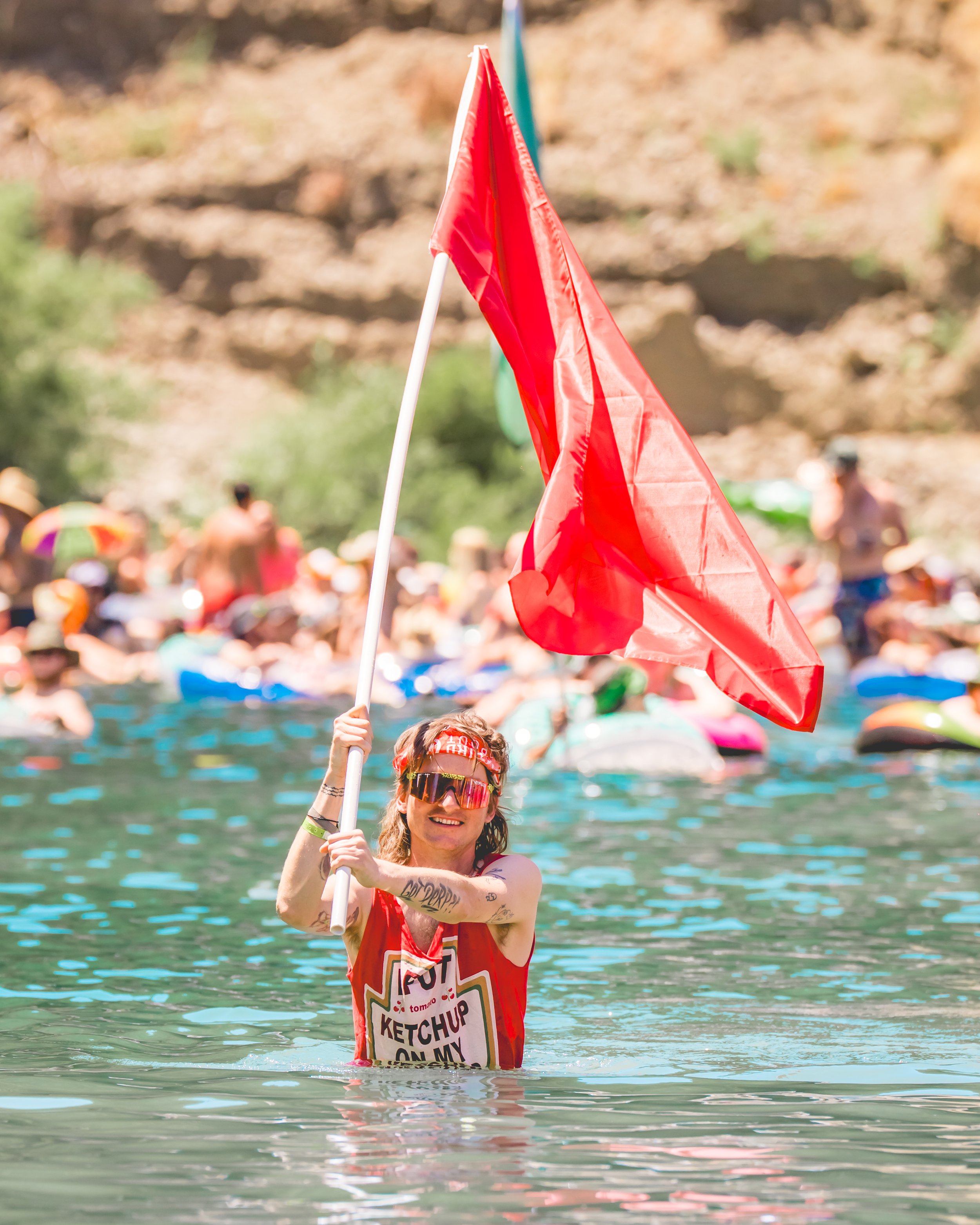 A woman in sunglasses and a red tank top holding a red flag in a water body during a large outdoor event with many people in the background.