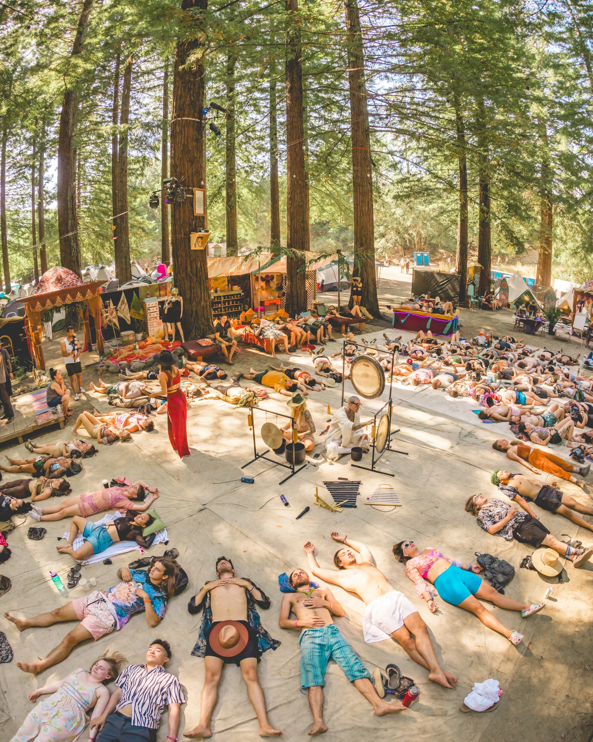 Group of people lying on the ground in a forest, practicing meditation. In the center, two musicians are playing gongs, and others are sitting or standing nearby. The setting includes tents and booths among tall trees, suggesting an outdoor event or 