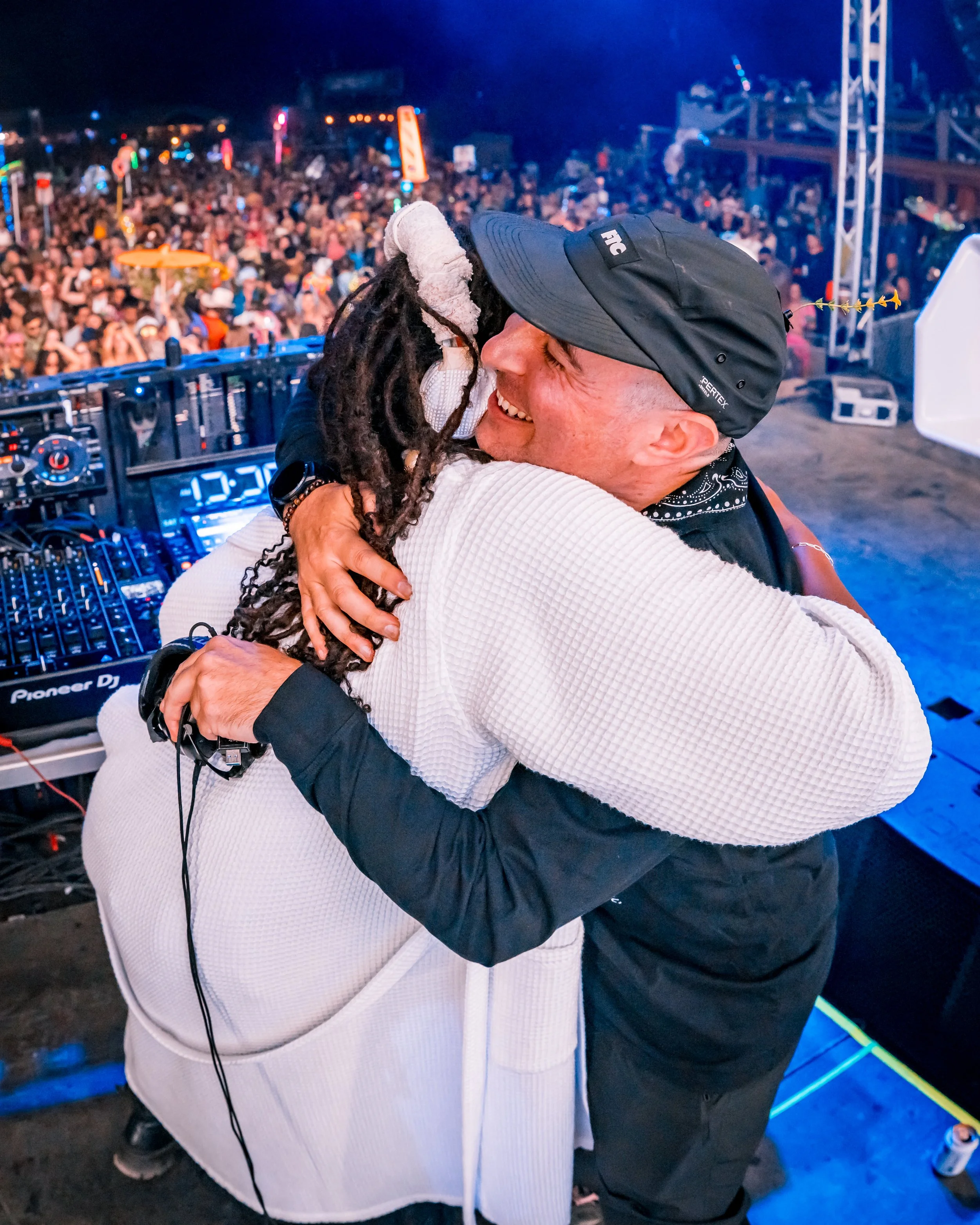 Two people embracing on a stage with a DJ setup, with a large crowd of people in the background at a lively concert or festival.