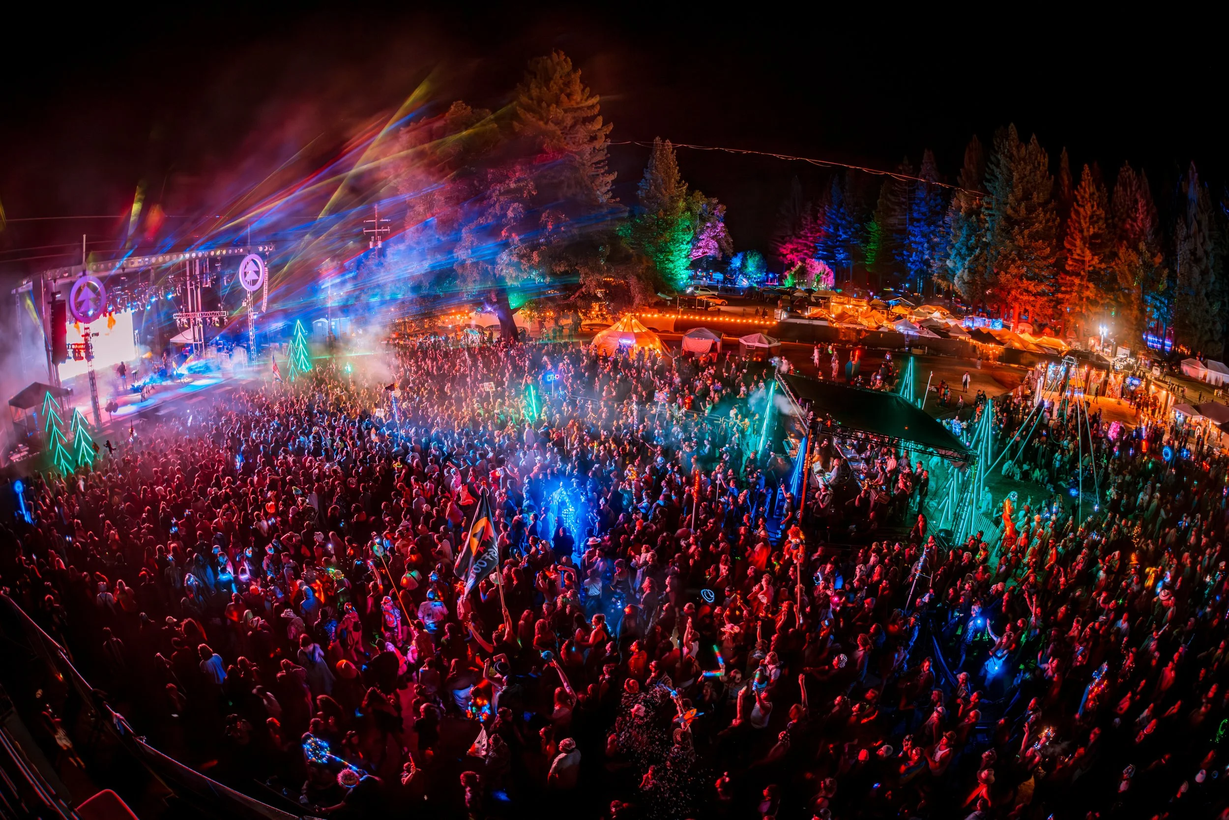 Nighttime outdoor concert with colorful lights, large crowd, and stage in the background, surrounded by trees lit with multicolored lights.