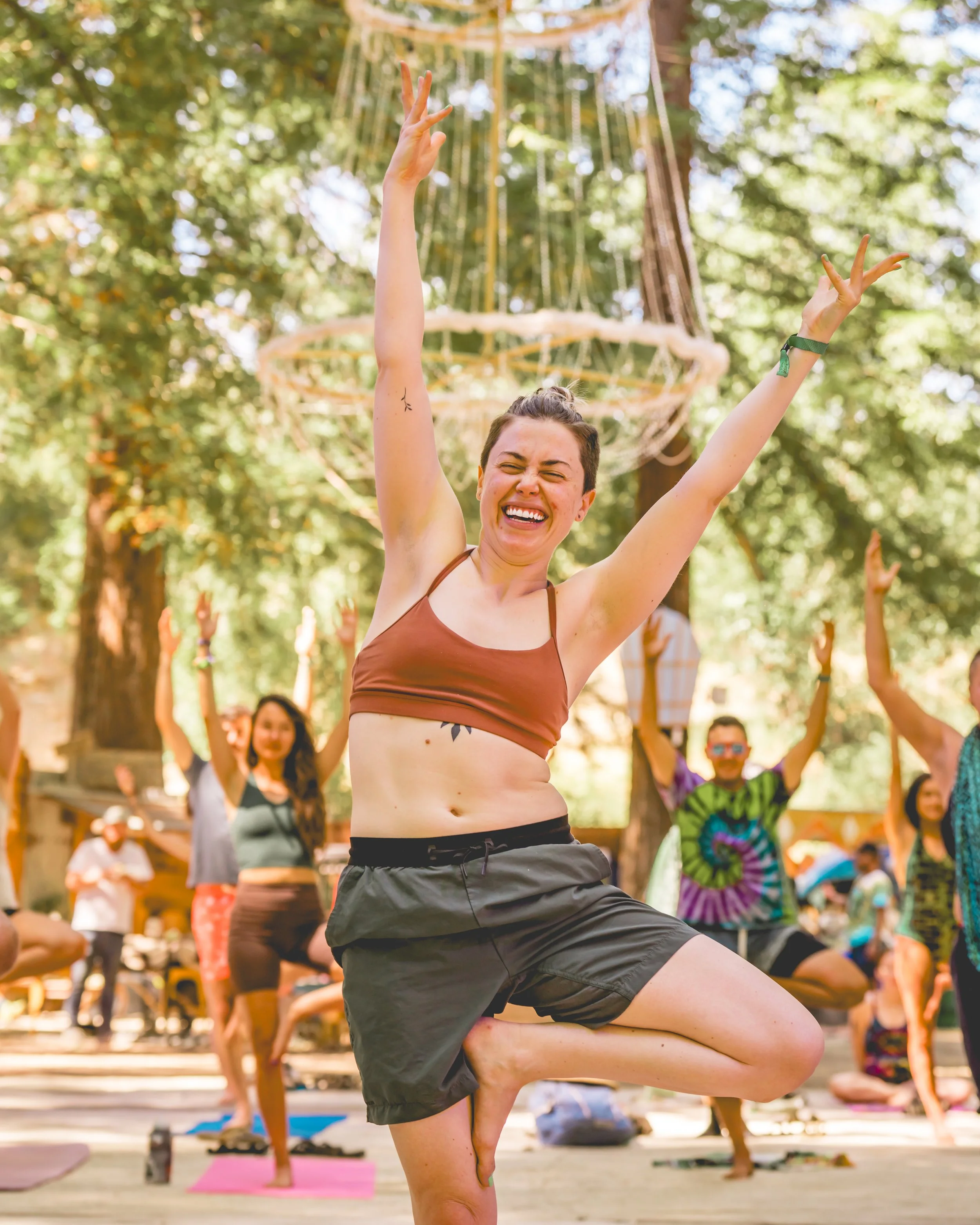 A woman with short hair doing a yoga pose at an outdoor yoga class surrounded by other participants, with trees and a wooden structure in the background.