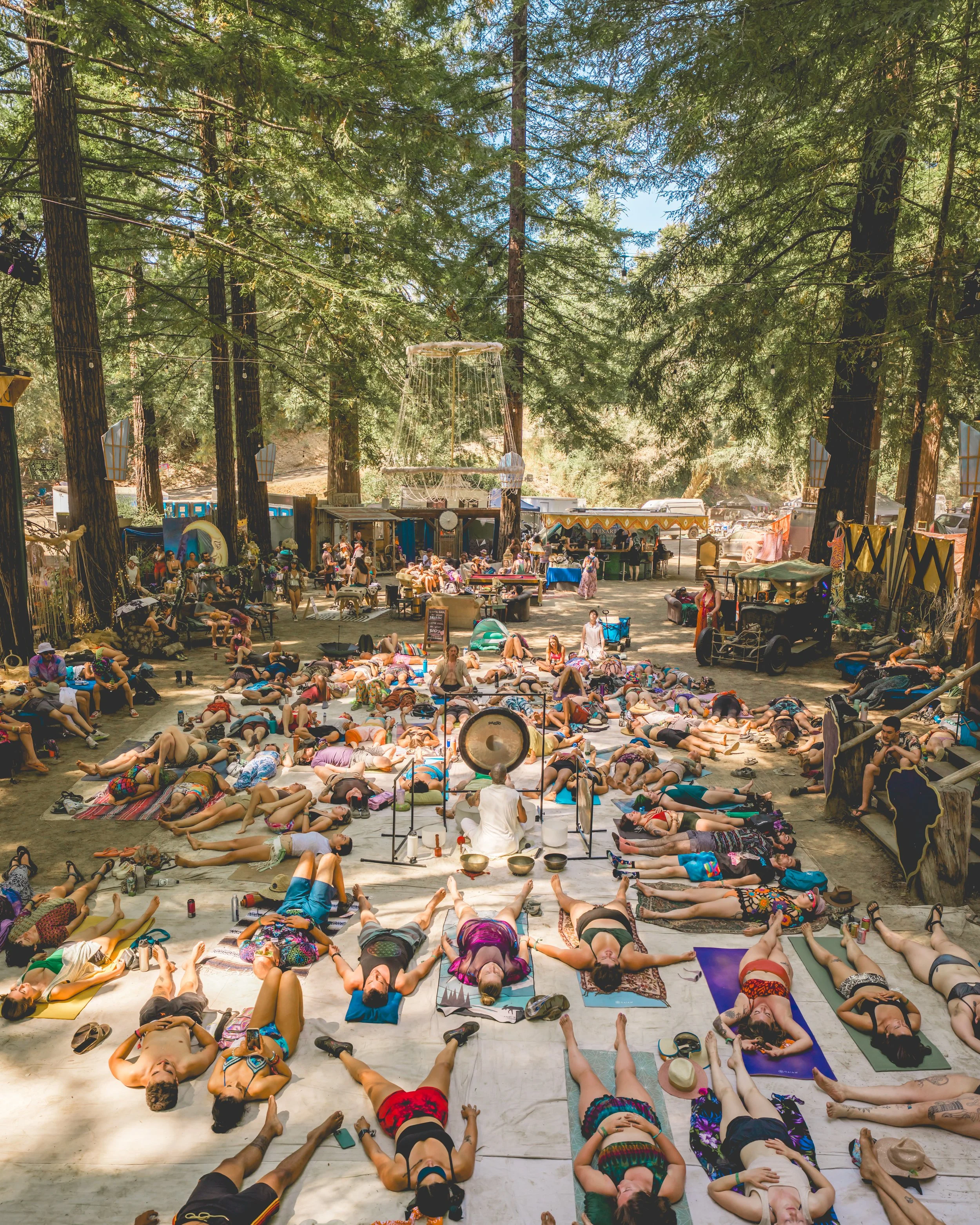 Large group of people practicing yoga outdoors in a forest, lying on mats with a large gong in the center, surrounded by trees, tents, and vendor booths.