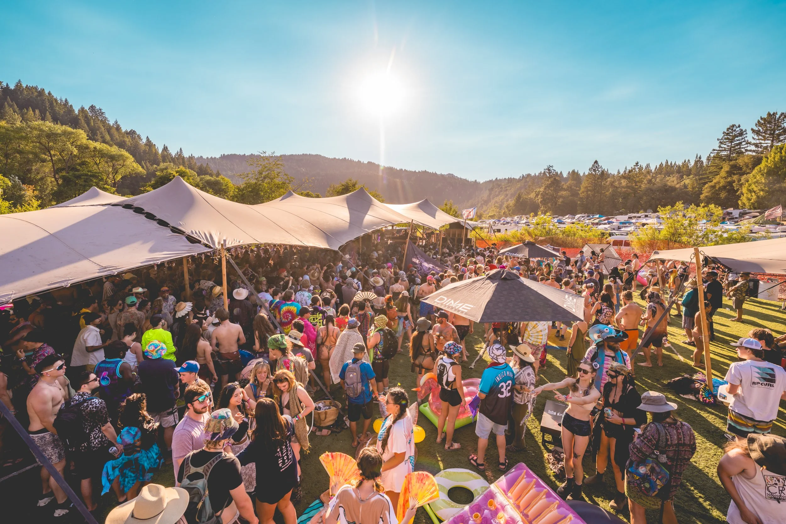 Crowd of people at outdoor festival with tents and umbrellas on a sunny day, surrounded by trees and mountains in the background.