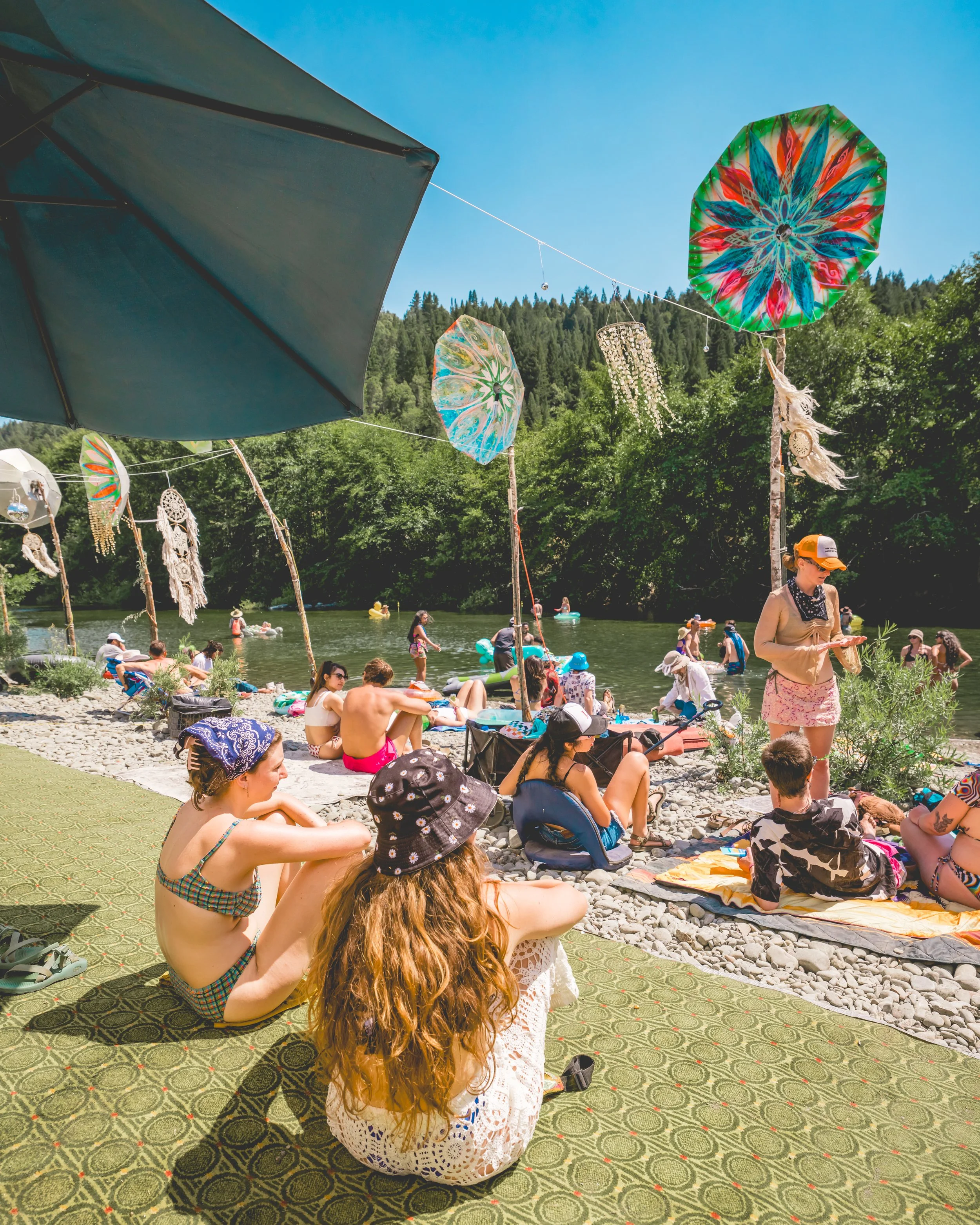 People relaxing on a pebbled riverside, with colorful umbrellas and decorative hanging ornaments, surrounded by green trees and mountains under a clear blue sky.