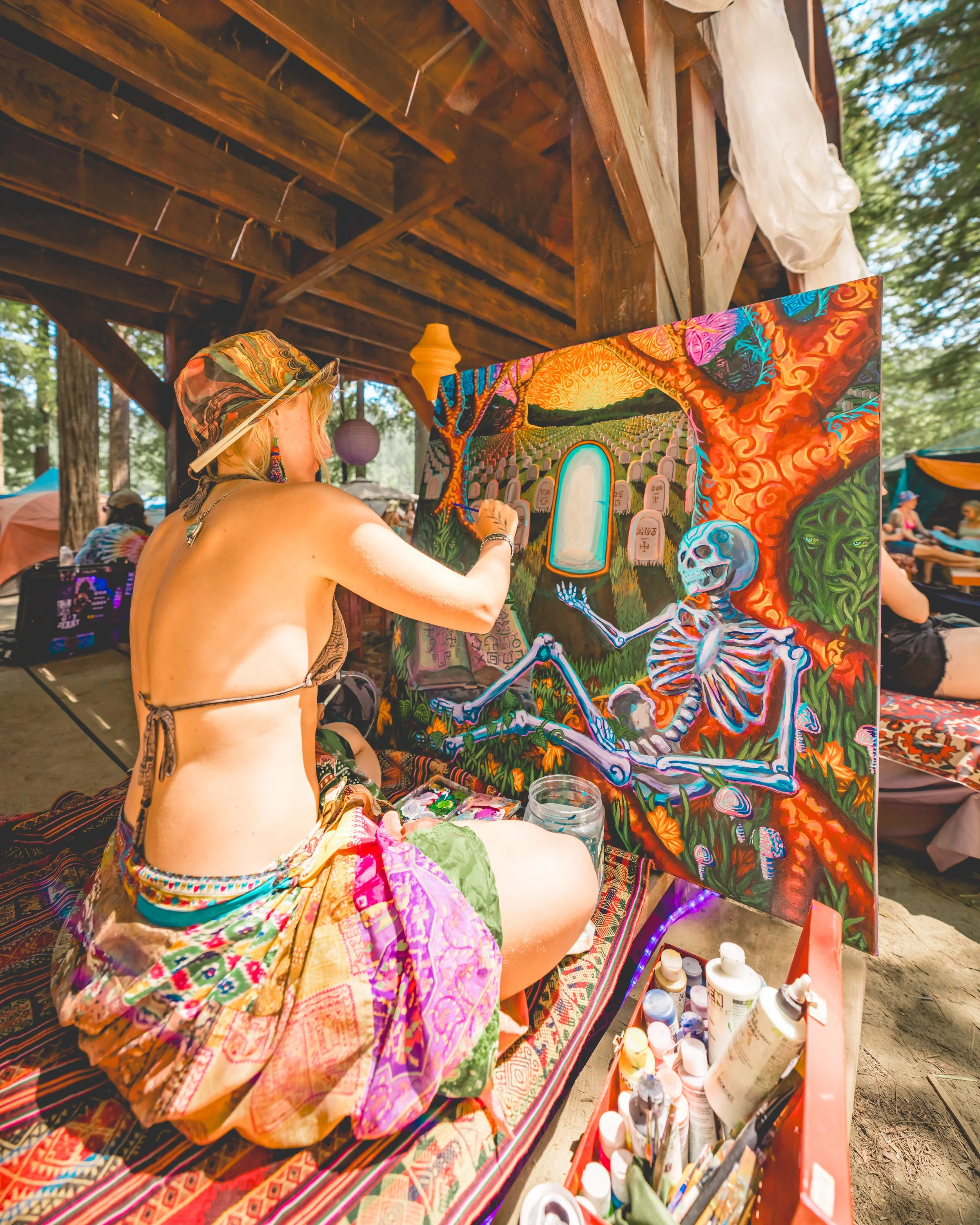 A woman in bohemian clothing and a hat painting a vibrant, surreal painting featuring a skeleton, gravestones, and fiery trees outdoors under a wooden canopy.