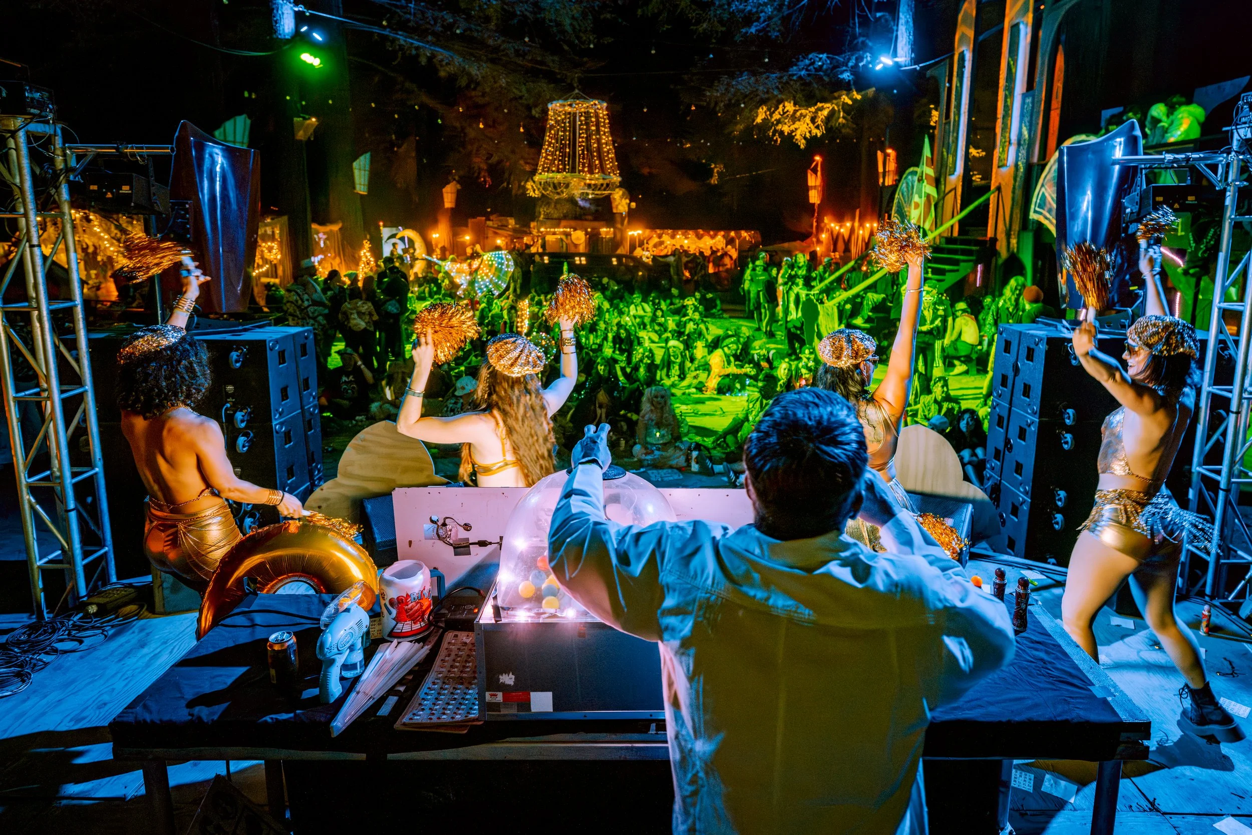 View of a lively outdoor dance party at night with dancers wearing colorful costumes and pom-poms, illuminated by vibrant green, yellow, and orange lights, with a crowd dancing in the background.