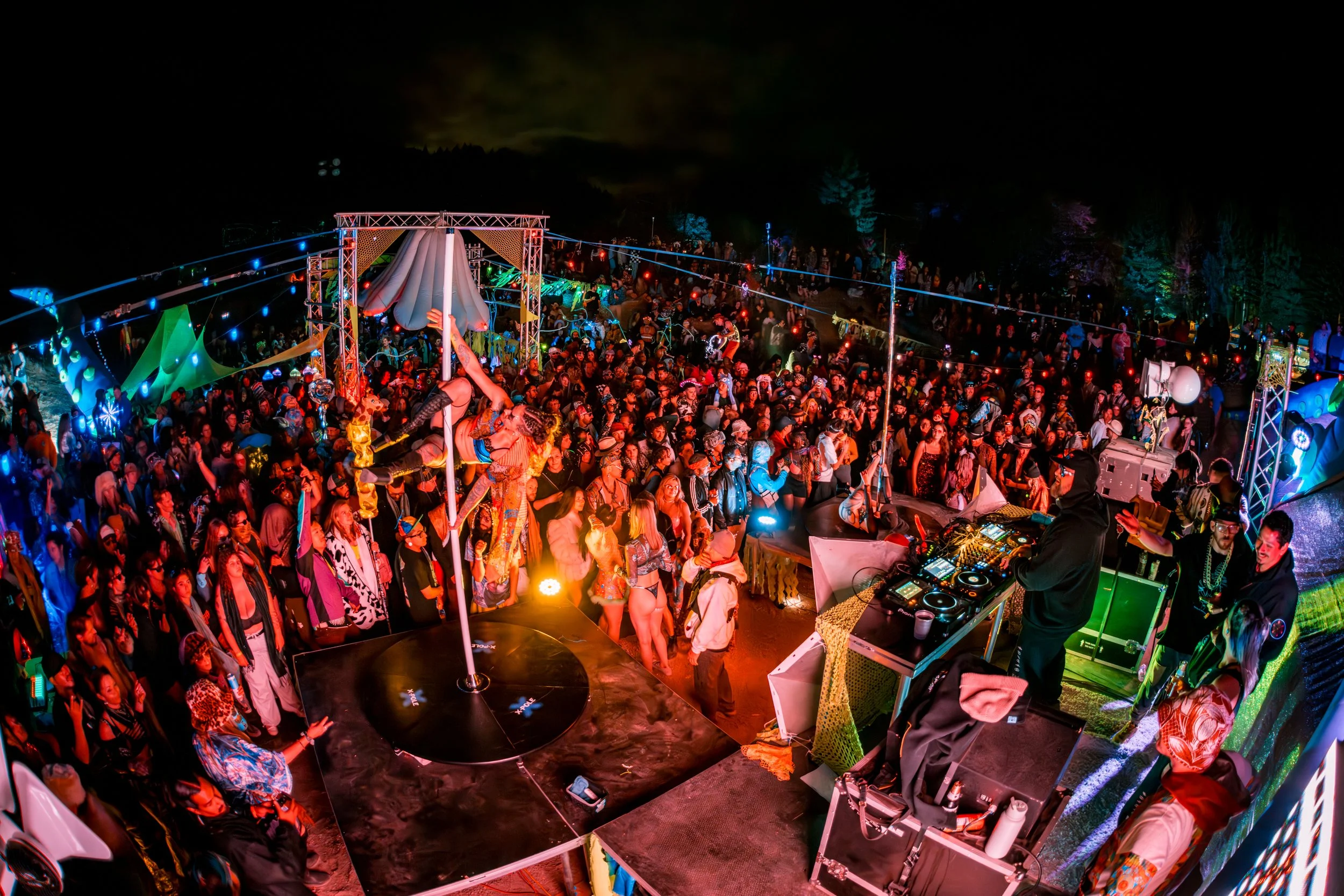 Nighttime music festival with a large crowd of people dancing and enjoying a stage performance with colorful lights. A DJ is performing on an elevated platform with equipment.