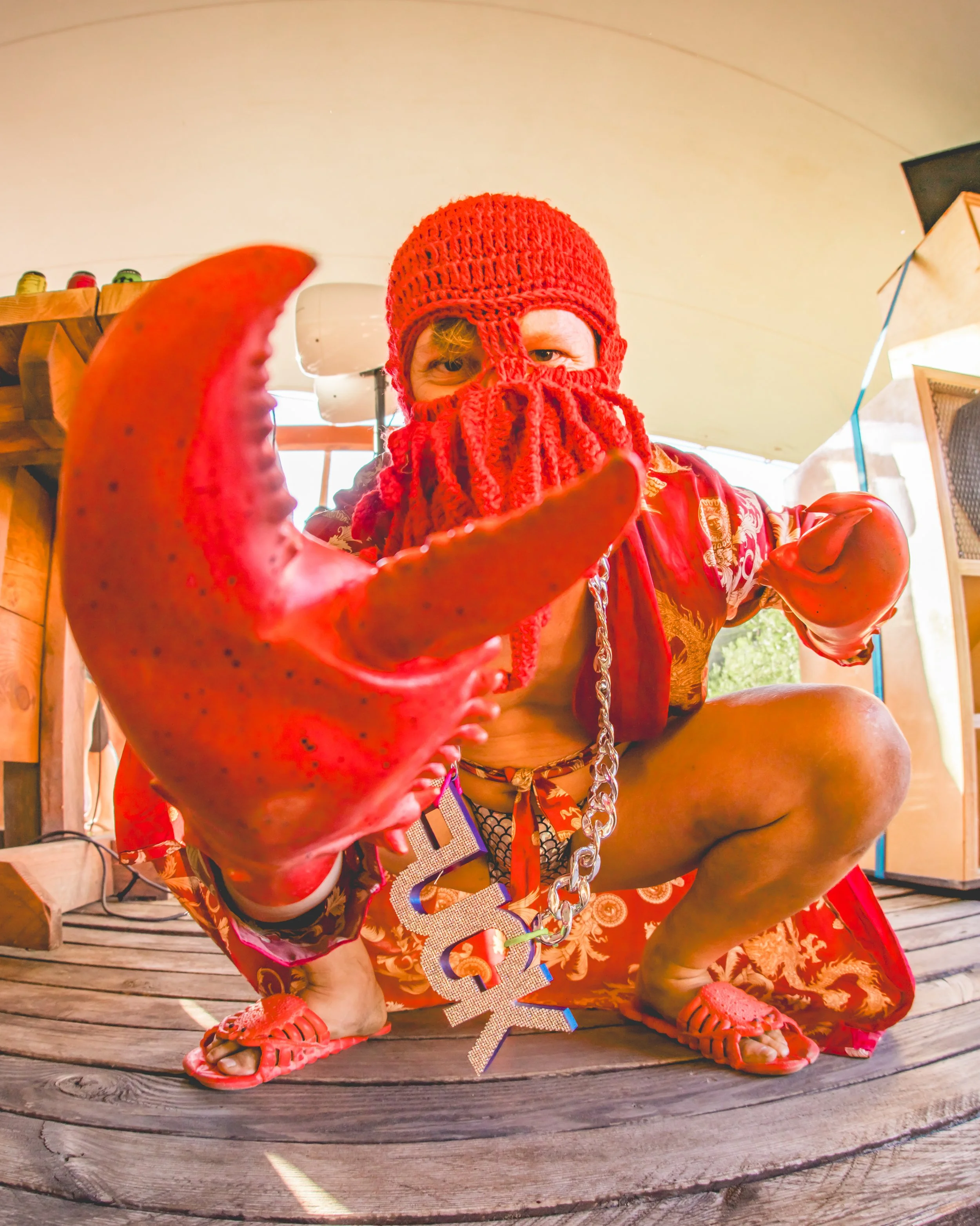 Person dressed in a red knit hat, beard, gloves, and slippers, holding a large lobster claw, inside a tent or outdoor structure.