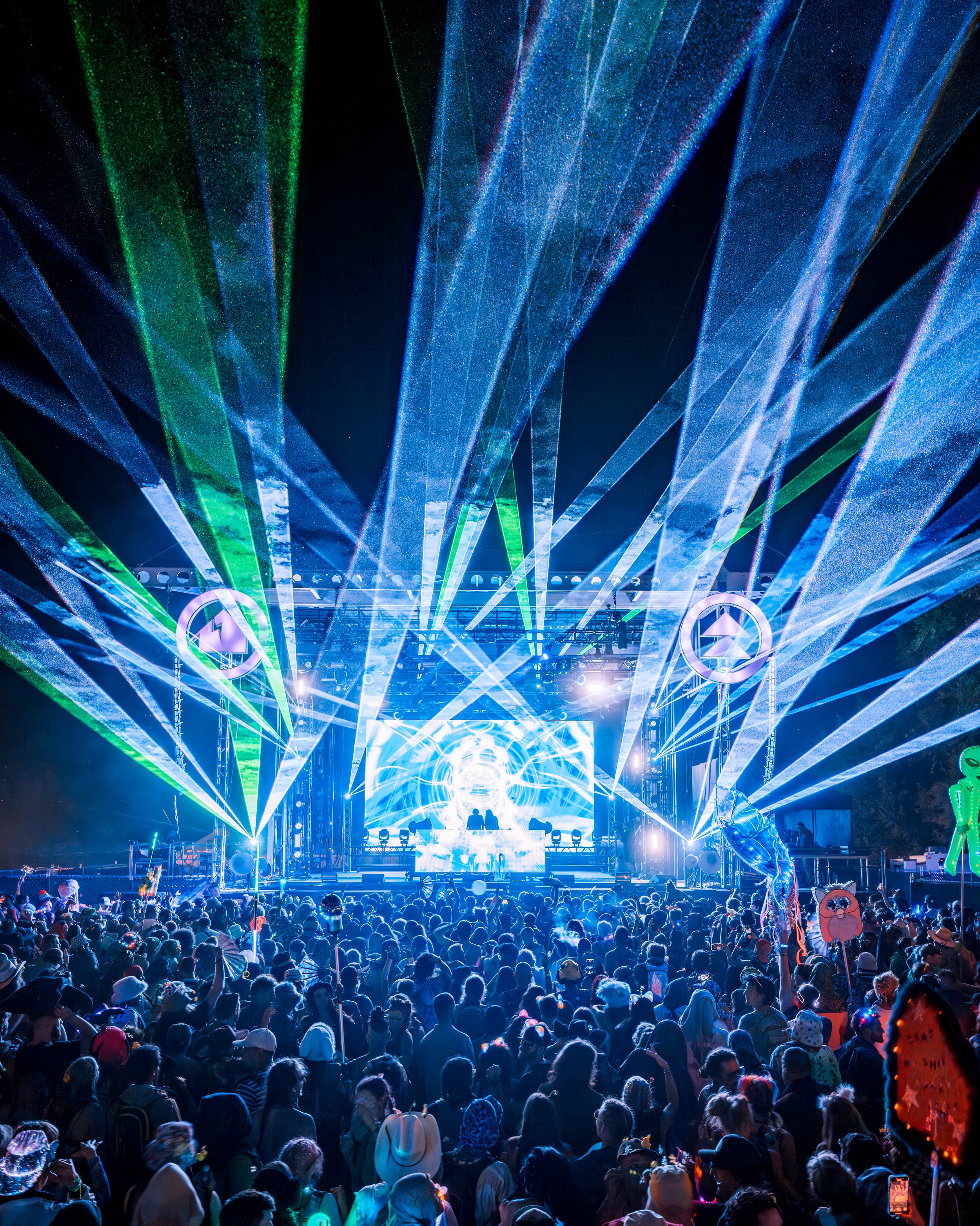 Crowd at a nighttime outdoor electronic music concert with bright laser lights and a DJ on stage.