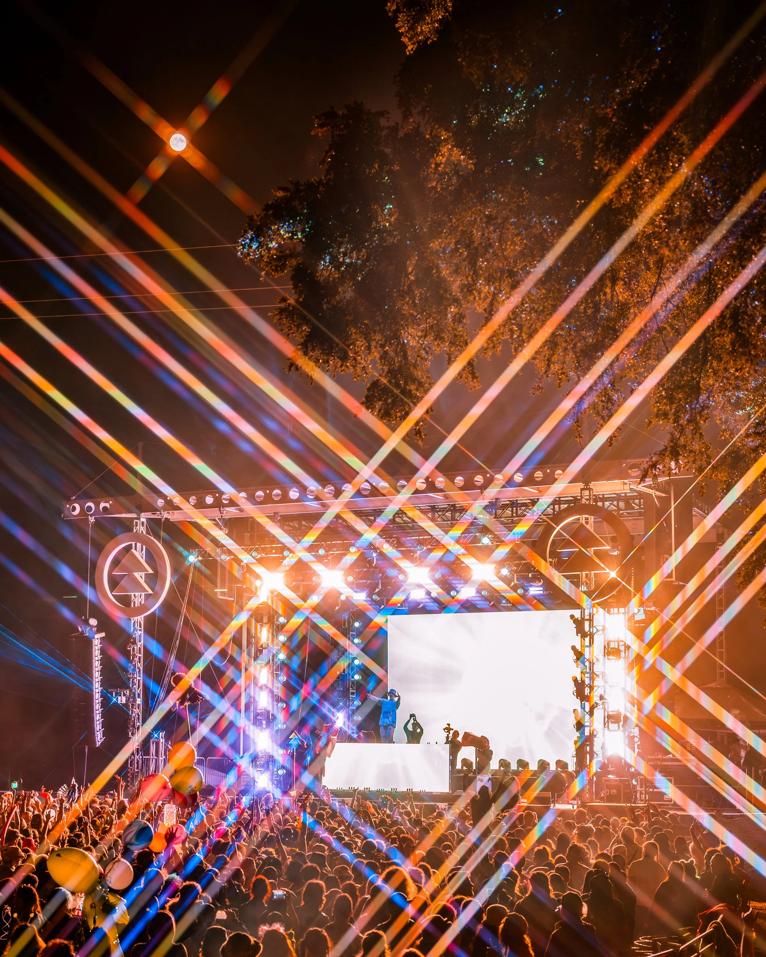 A concert stage at night with bright lights, a large crowd, balloons, and a backdrop with trees and a full moon overhead.