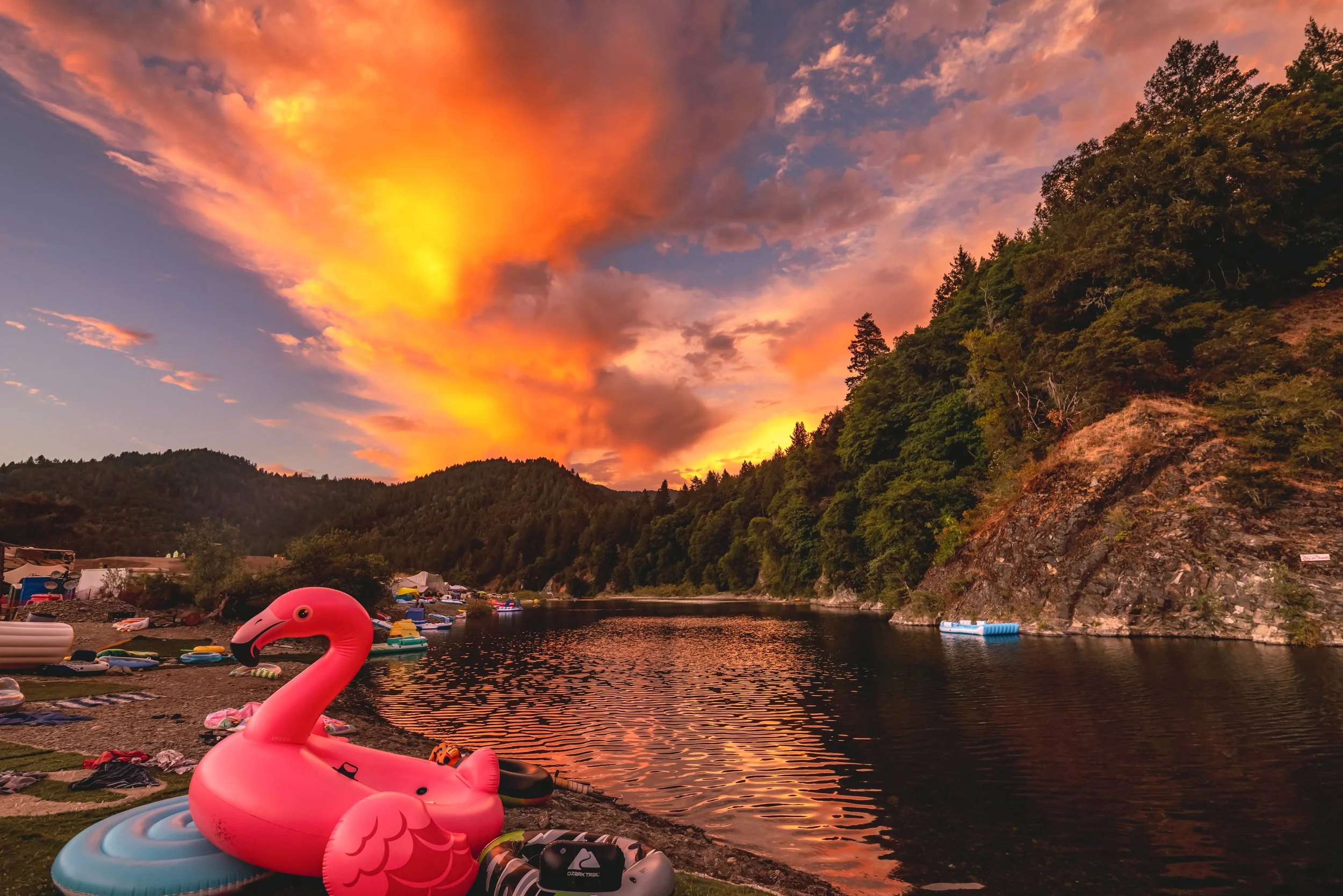 A lakeside scene at sunset with vibrant orange and pink clouds, a calm water body reflecting the sky, and a pink inflatable flamingo float on the shore surrounded by assorted watercraft and belongings.
