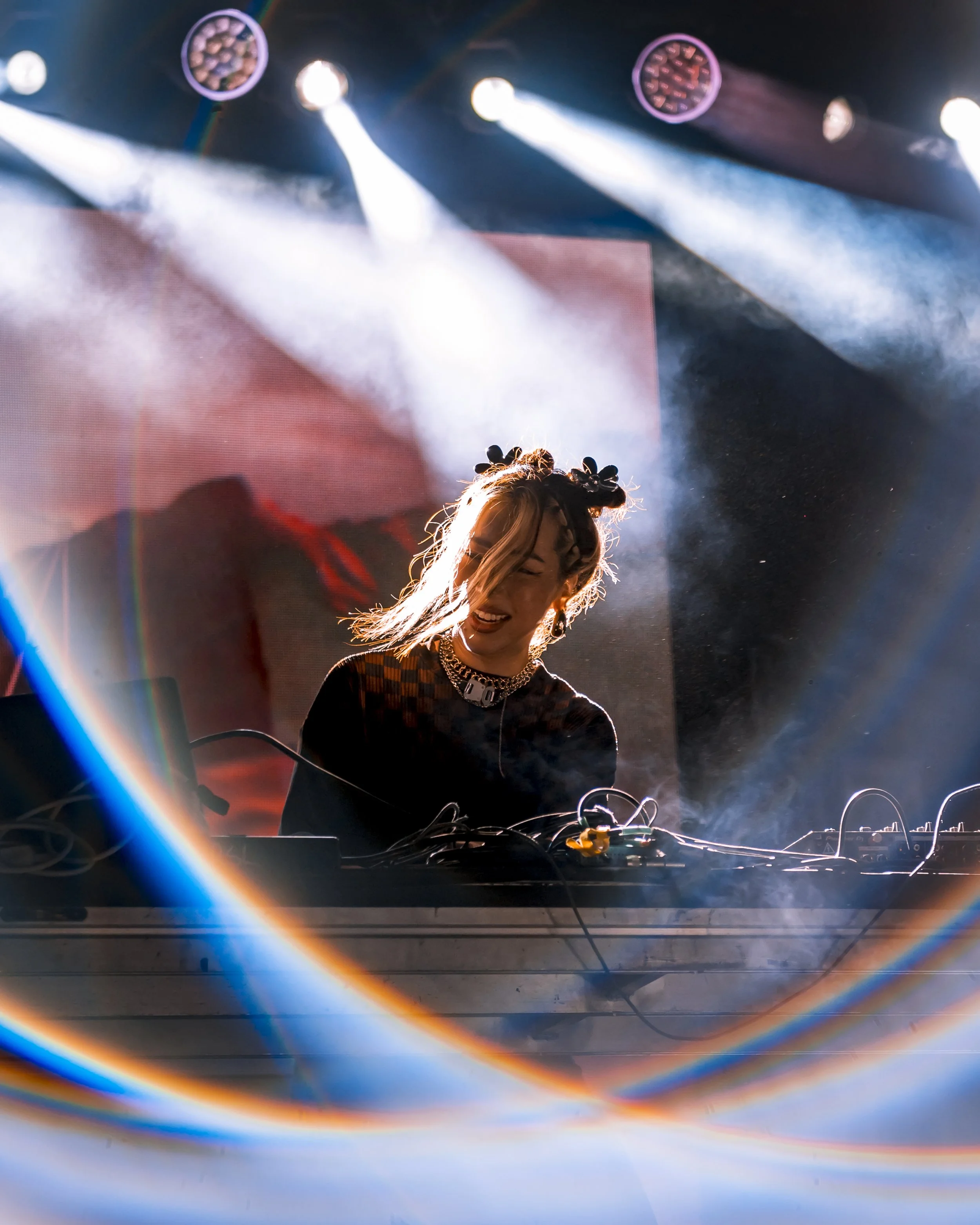 Female DJ performing on stage with colorful lights and smoke, wearing a headband and jewelry.