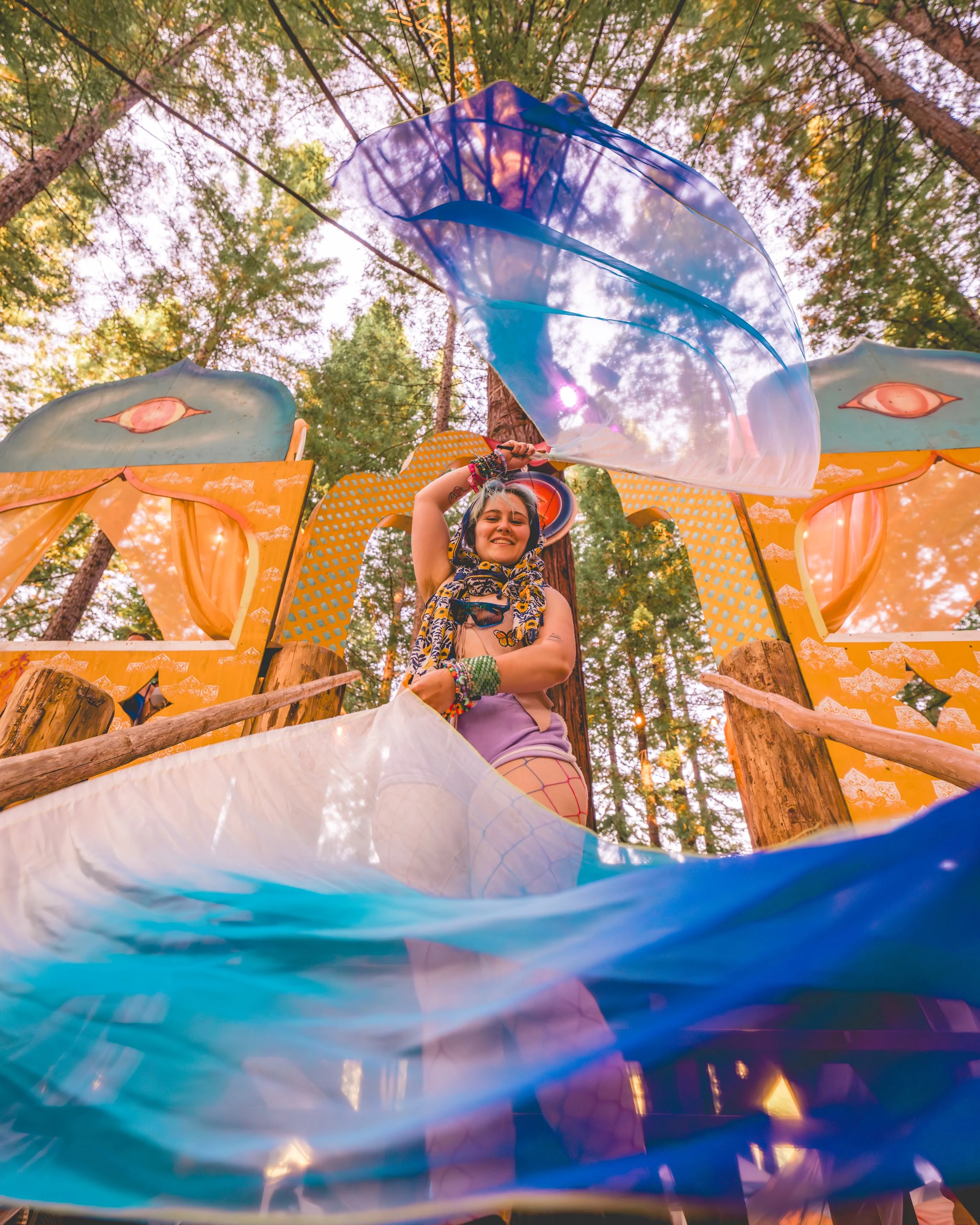 A smiling woman with colorful accessories, including bracelets and fishnet stockings, stands in front of a vibrant, fairy-tale-like outdoor setting with decorated structures and tall trees, holding a shiny blue kite.