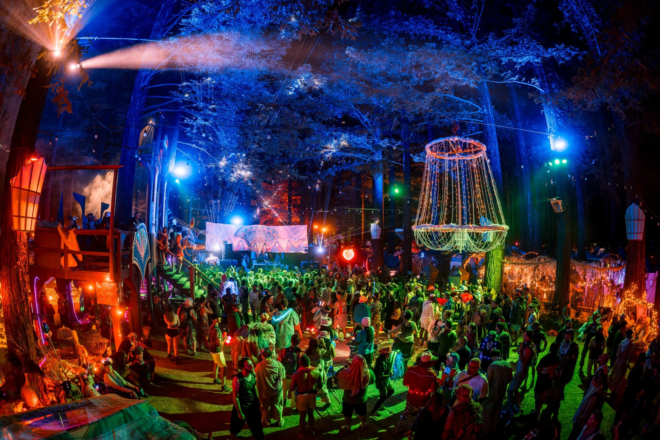 A vibrant nighttime scene at an outdoor festival in a forest, with colorful lights, a large crowd of people, illuminated decorations, and a stage with performers.
