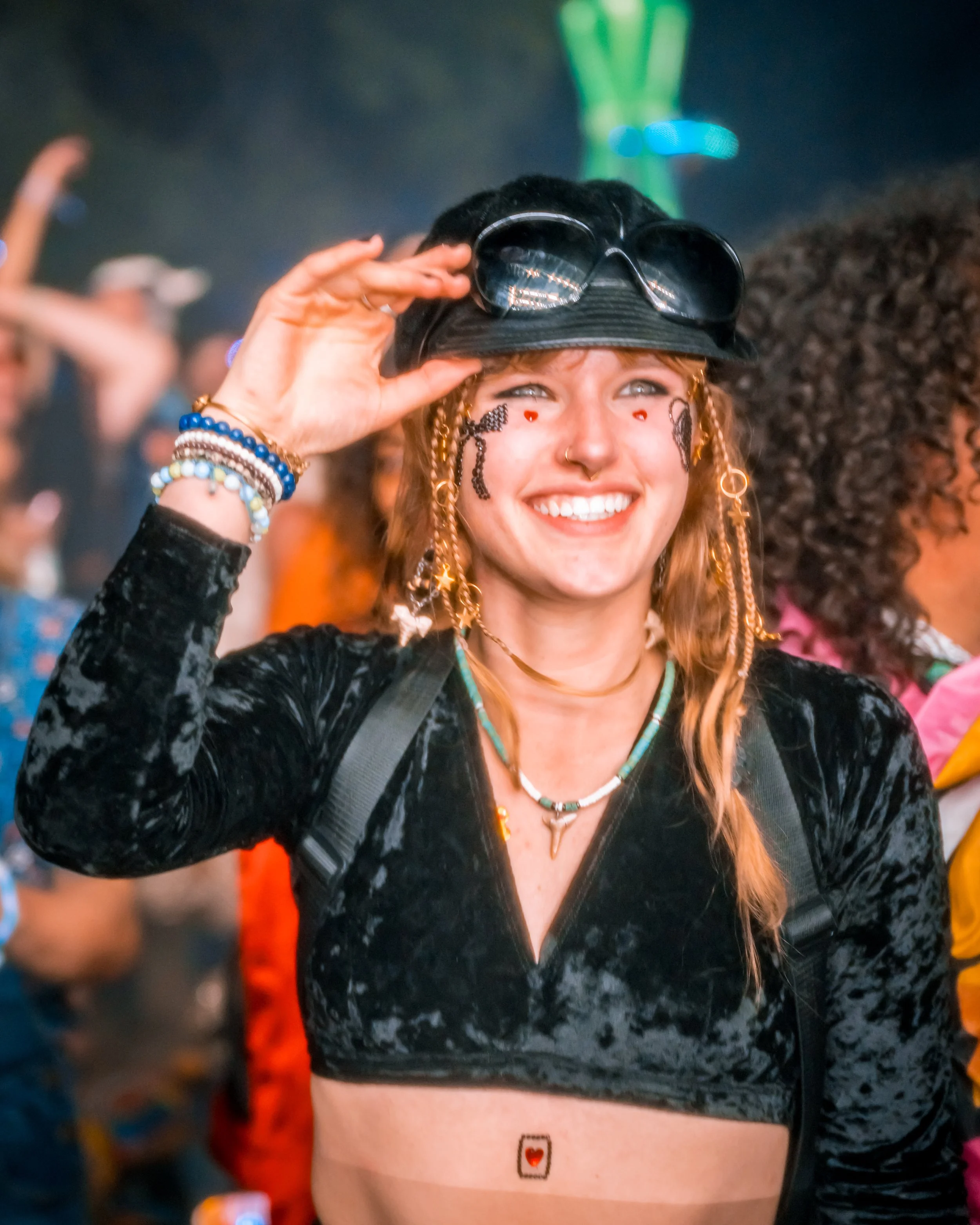 A smiling woman at a crowded event, wearing a black velvet crop top, jewelry, and accessories, and holding sunglasses on her head.
