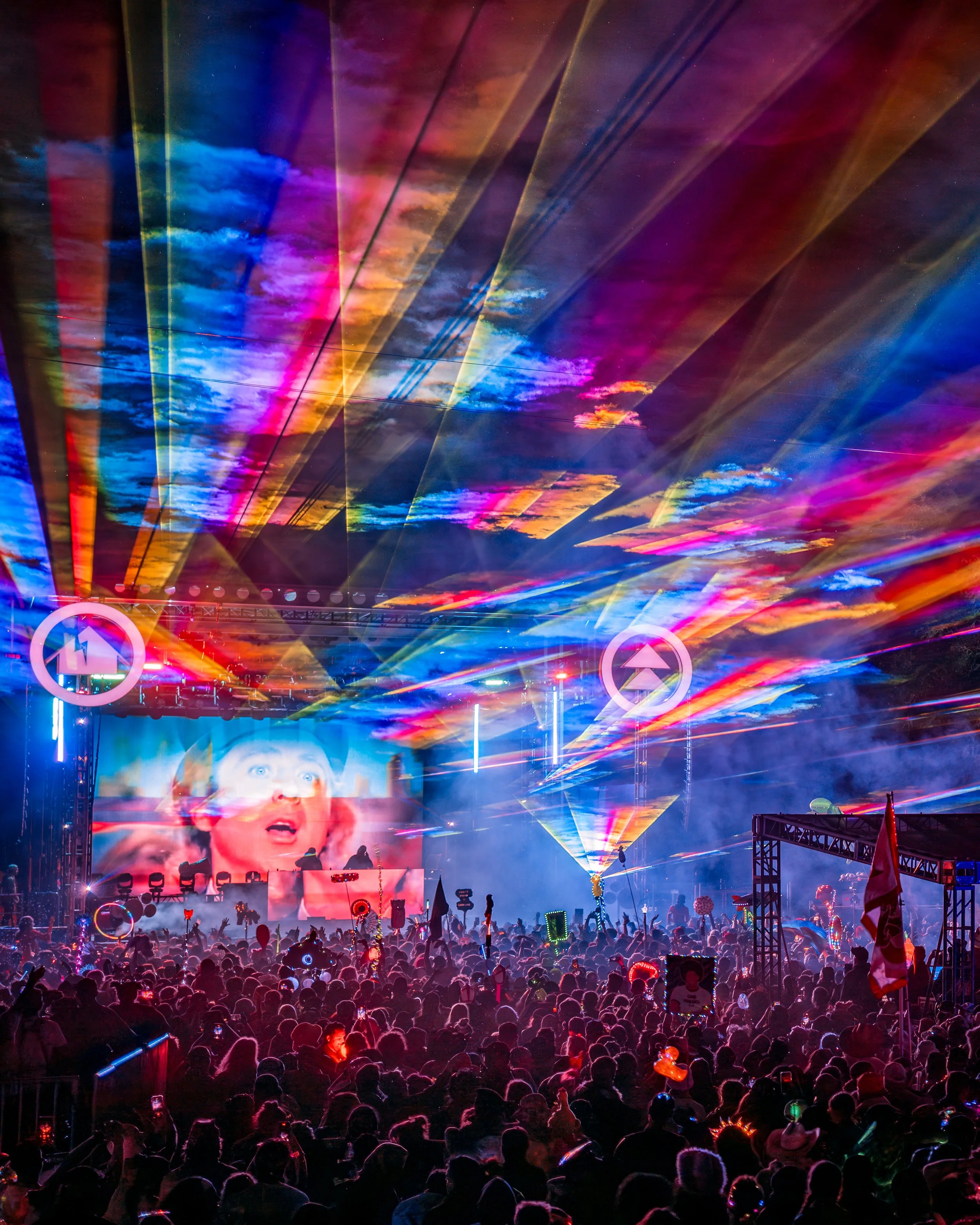 A crowded concert or festival event with colorful laser lights and a large screen displaying an excited woman, with visible stage equipment and attendees holding glow sticks.