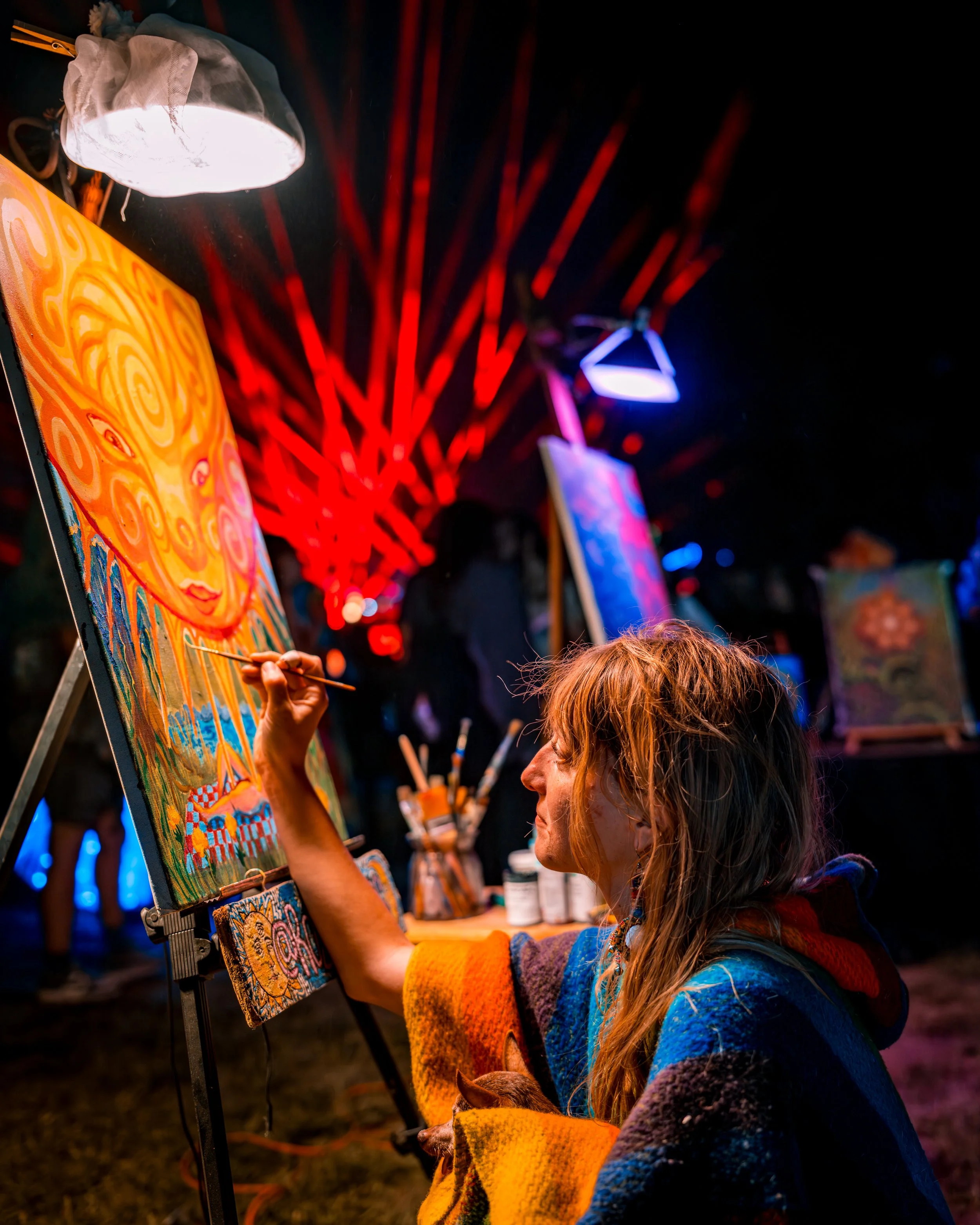 A woman painting a colorful abstract sun with a face on a canvas at a vibrant art event at night, with artistic lighting and other paintings in the background.