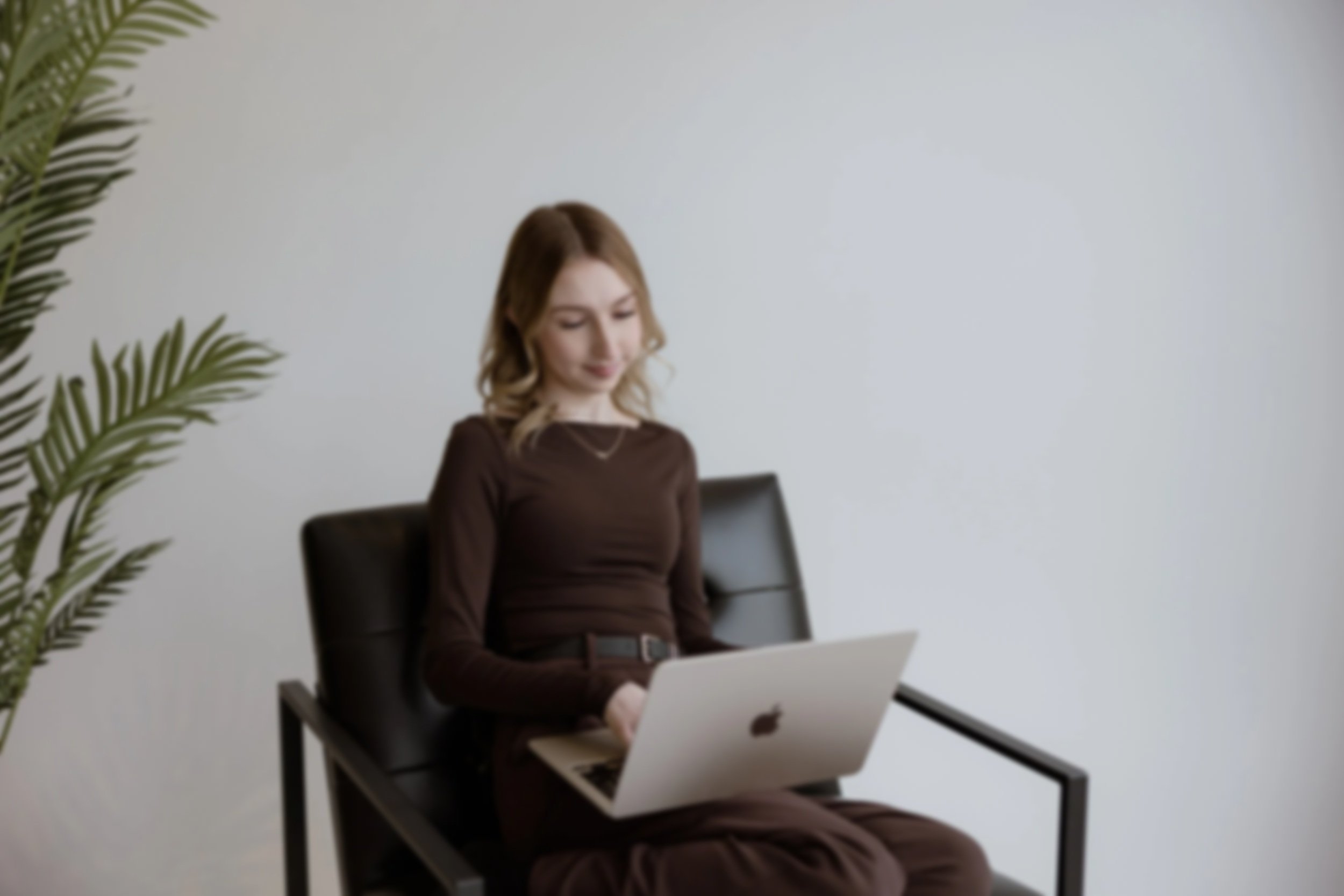 Woman sitting in a black chair using a silver MacBook laptop, with a green plant on the left and a plain white background.
