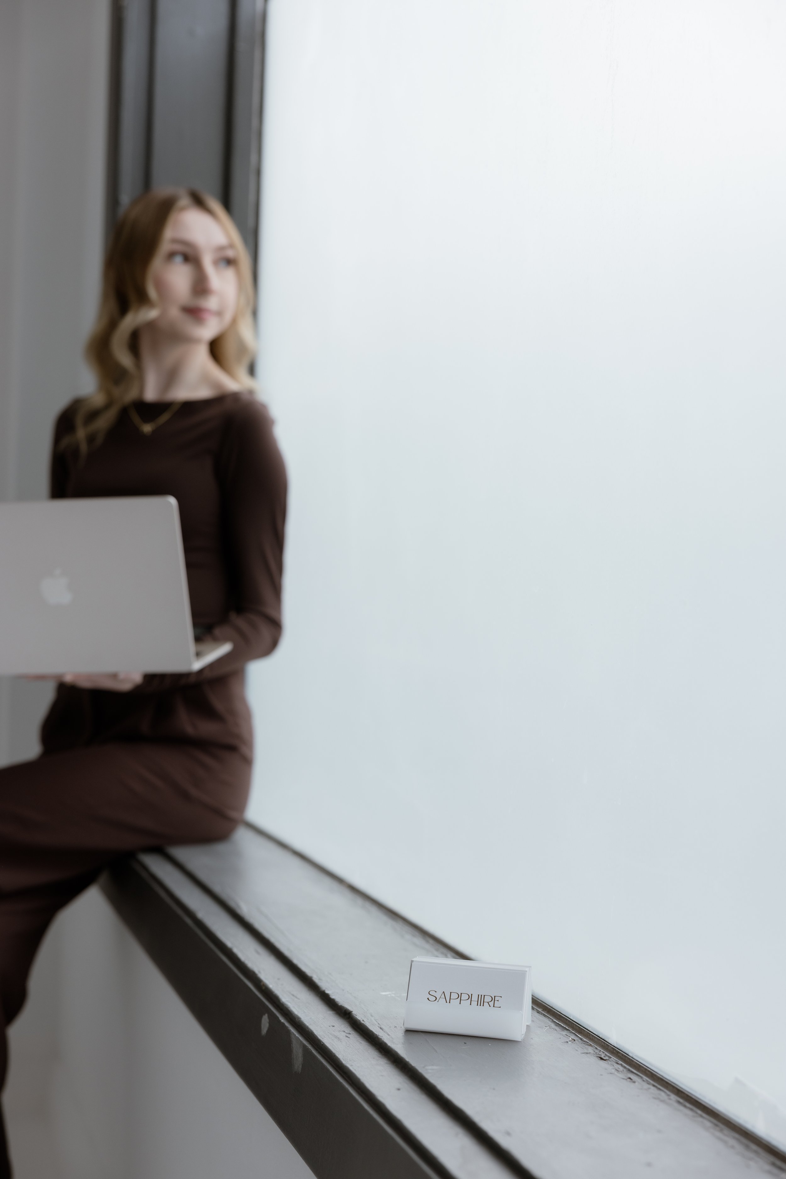 A woman with blonde hair sitting on a windowsill, holding a silver MacBook laptop, with a small white sign labeled "SAPPHIRE" on the windowsill.