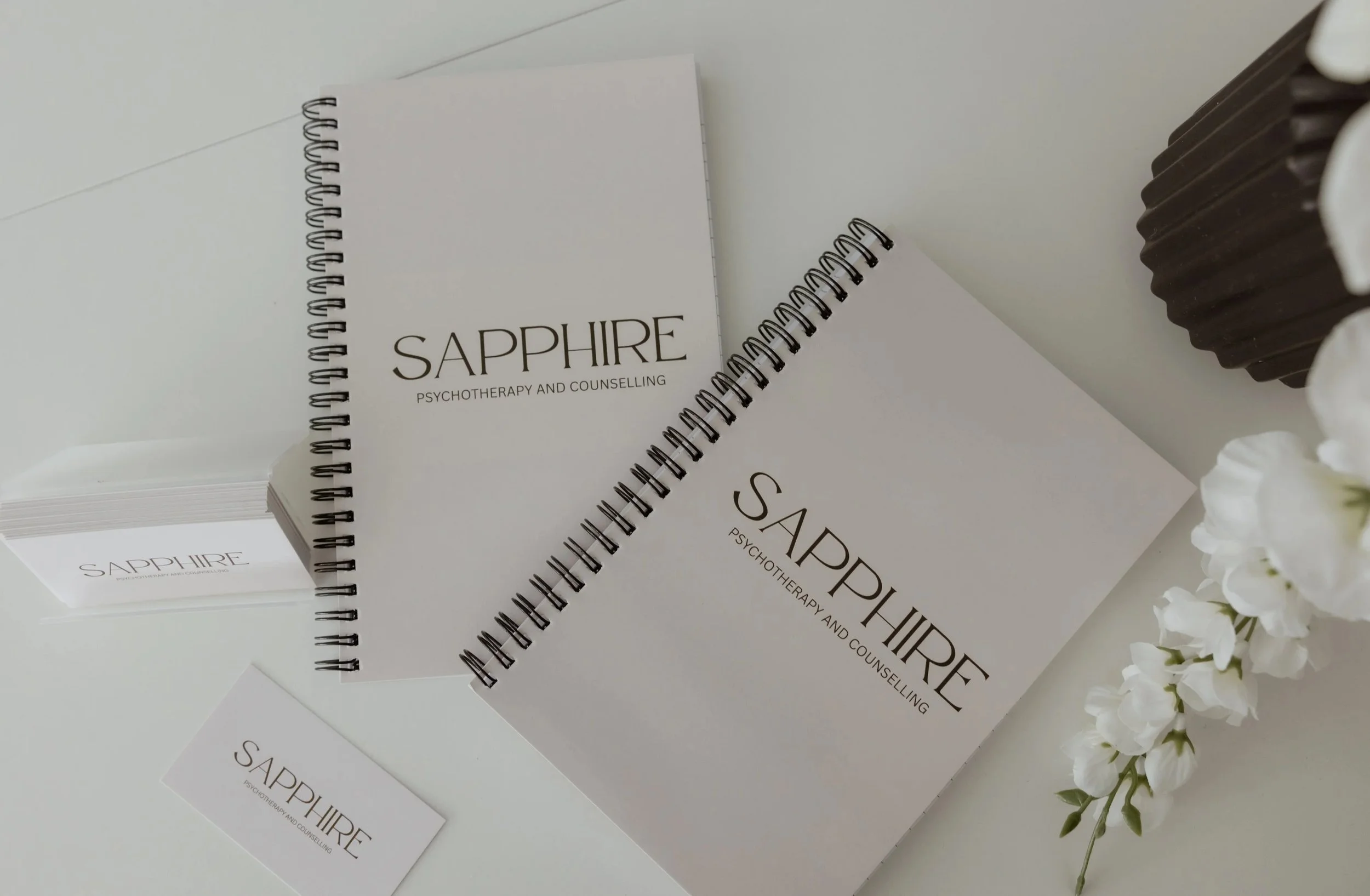 A collection of Sapphire psychotherapy and counseling notebooks, business cards, and a small flower arrangement on a white surface.