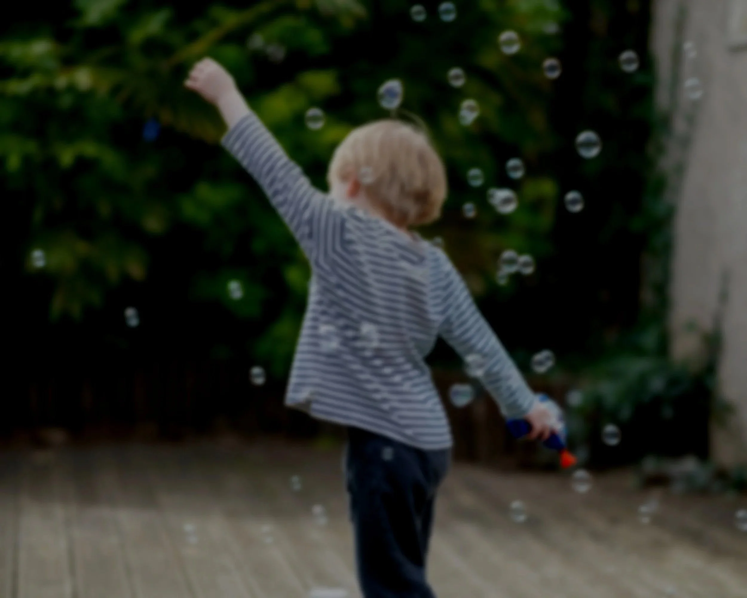 Child playing with bubbles outdoors on a wooden deck, with greenery in the background.