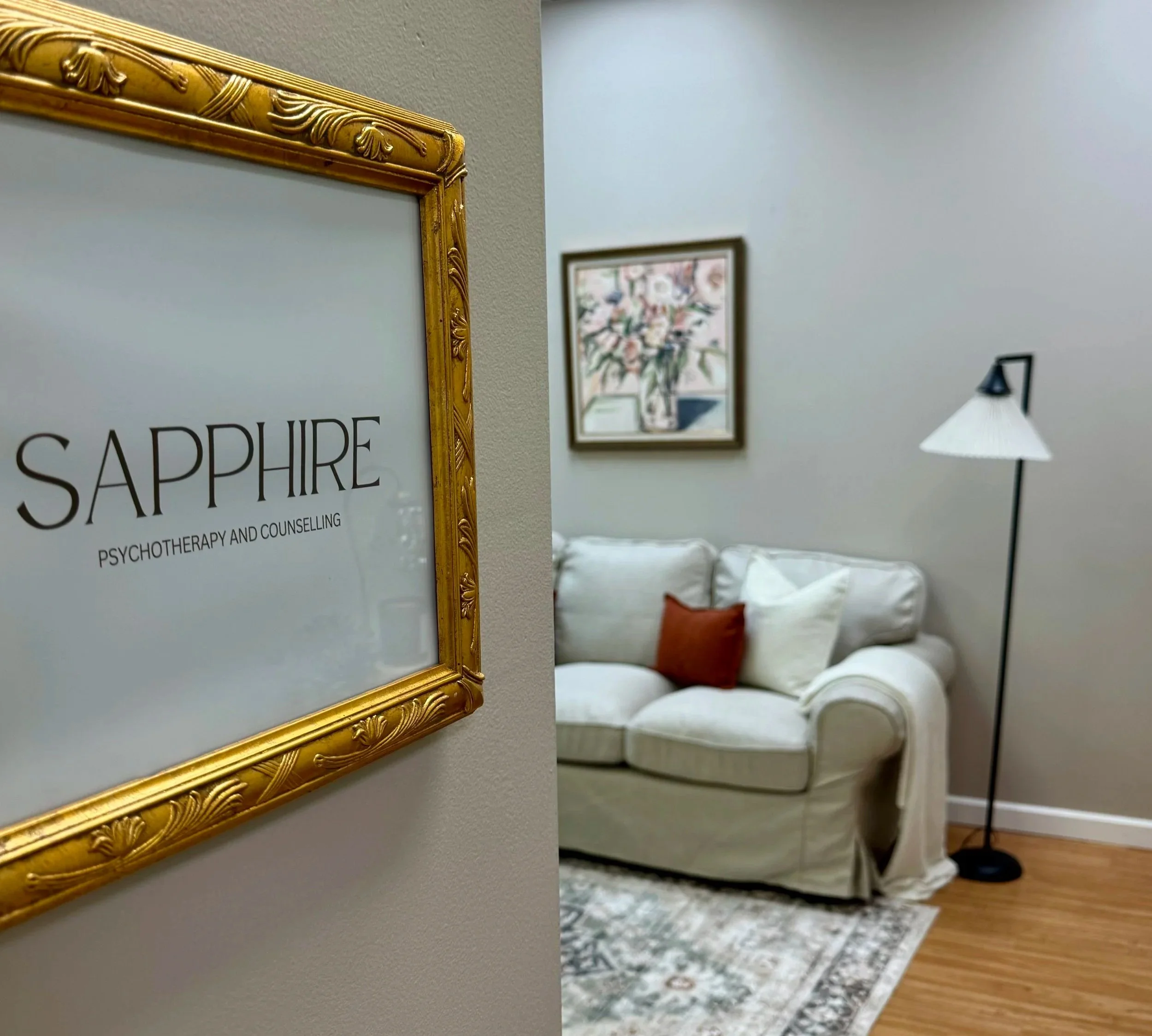 A sign that reads 'SAPPHIRE PSYCHOTHERAPY AND COUNSELLING' in a gold ornate frame, with a cozy room in the background featuring a white couch, decorative pillows, a framed floral painting, and a floor lamp.