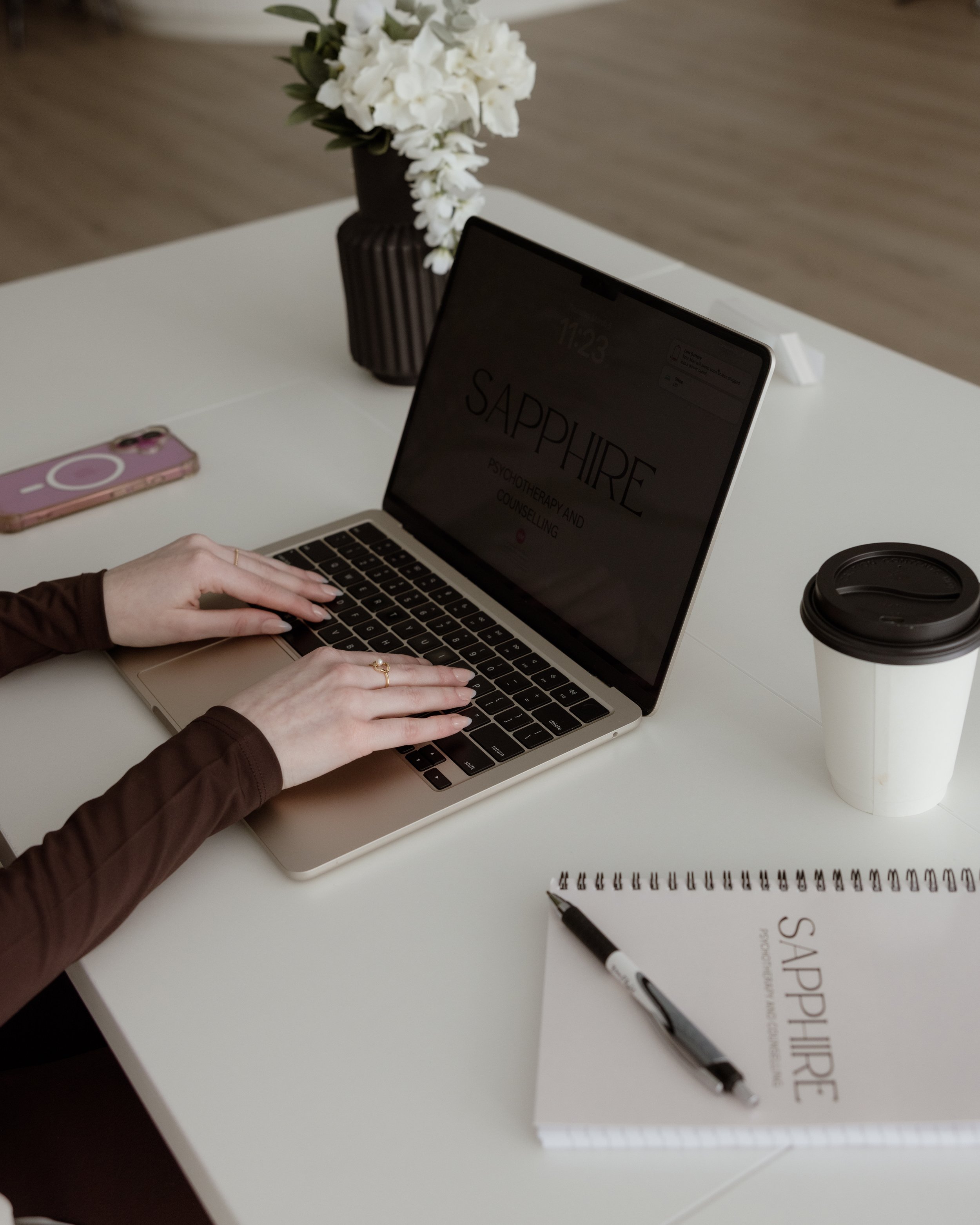 A person working on a laptop at a white desk with a notebook, pen, disposable coffee cup, pink phone, and a vase of white flowers.