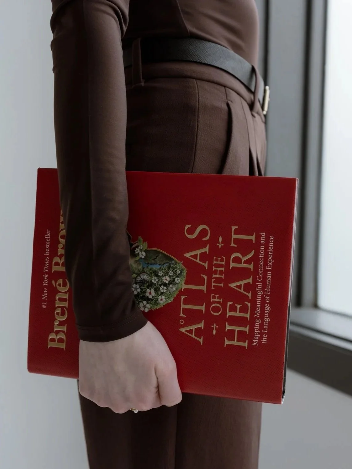 A person dressed in brown pants and a matching top holds a red book titled 'Atlas of the Heart' by Brené Brown, with a floral design on the cover, standing near a window.