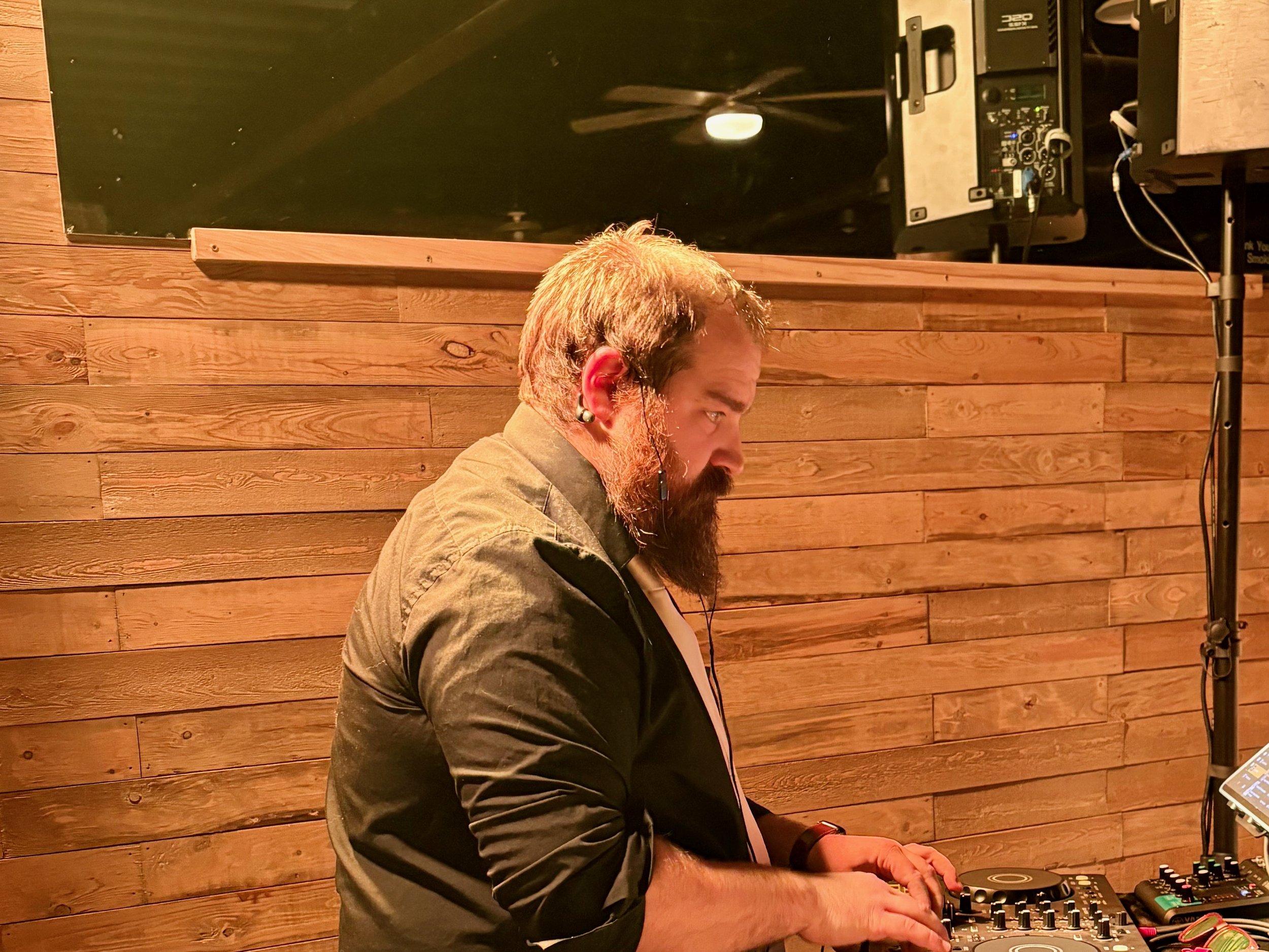 A man with a beard and mustache working as a DJ, wearing a black shirt and using DJ equipment in front of a wooden wall.