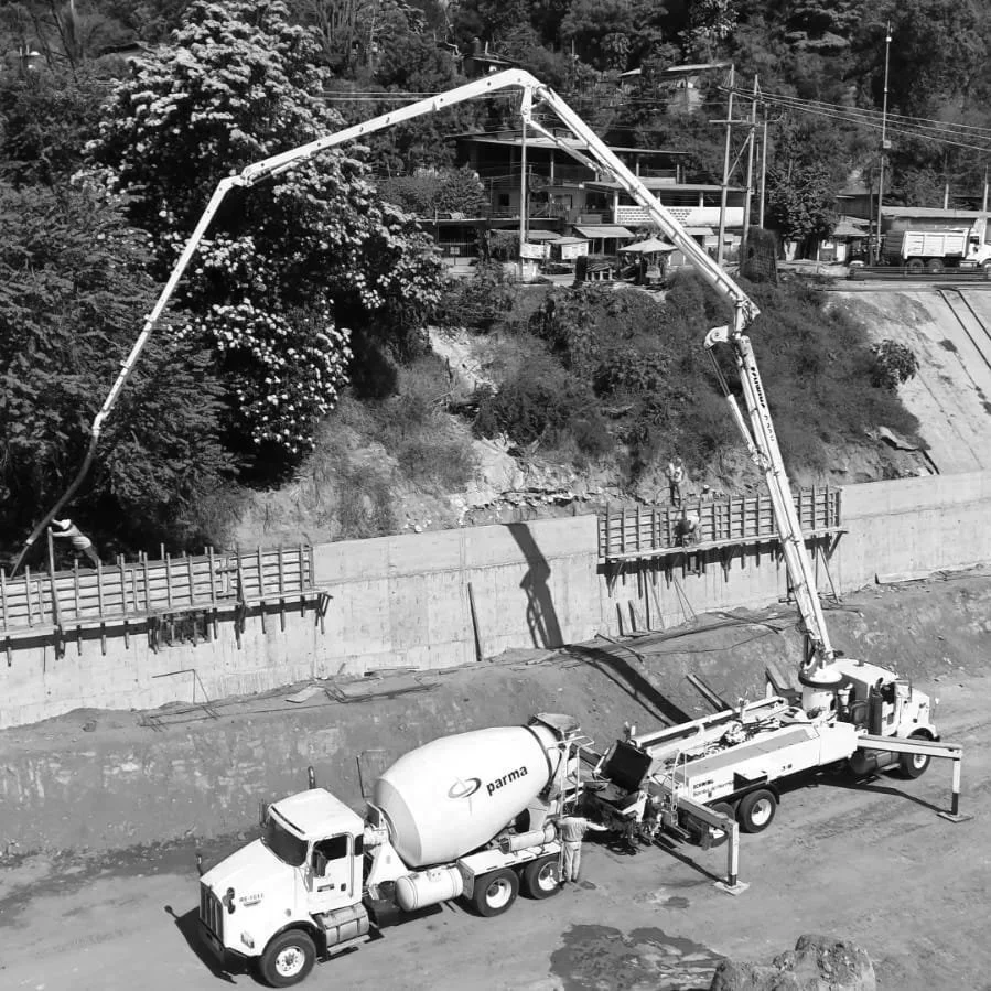 Camión bomba vertiendo concreto en una construcción en una ladera con casas y árboles en el fondo, en un ambiente de trabajo al aire libre.