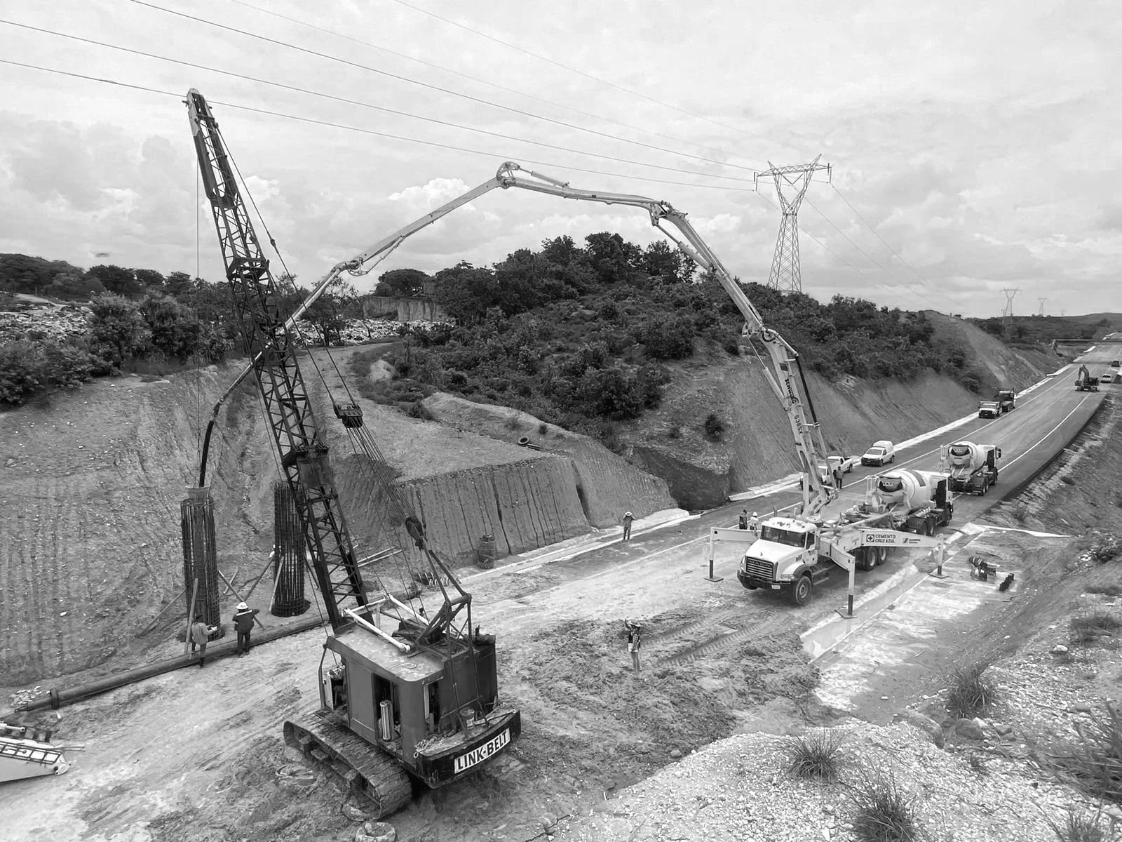 Construcción en una autopista con maquinaria y personal, con líneas eléctricas en el fondo.