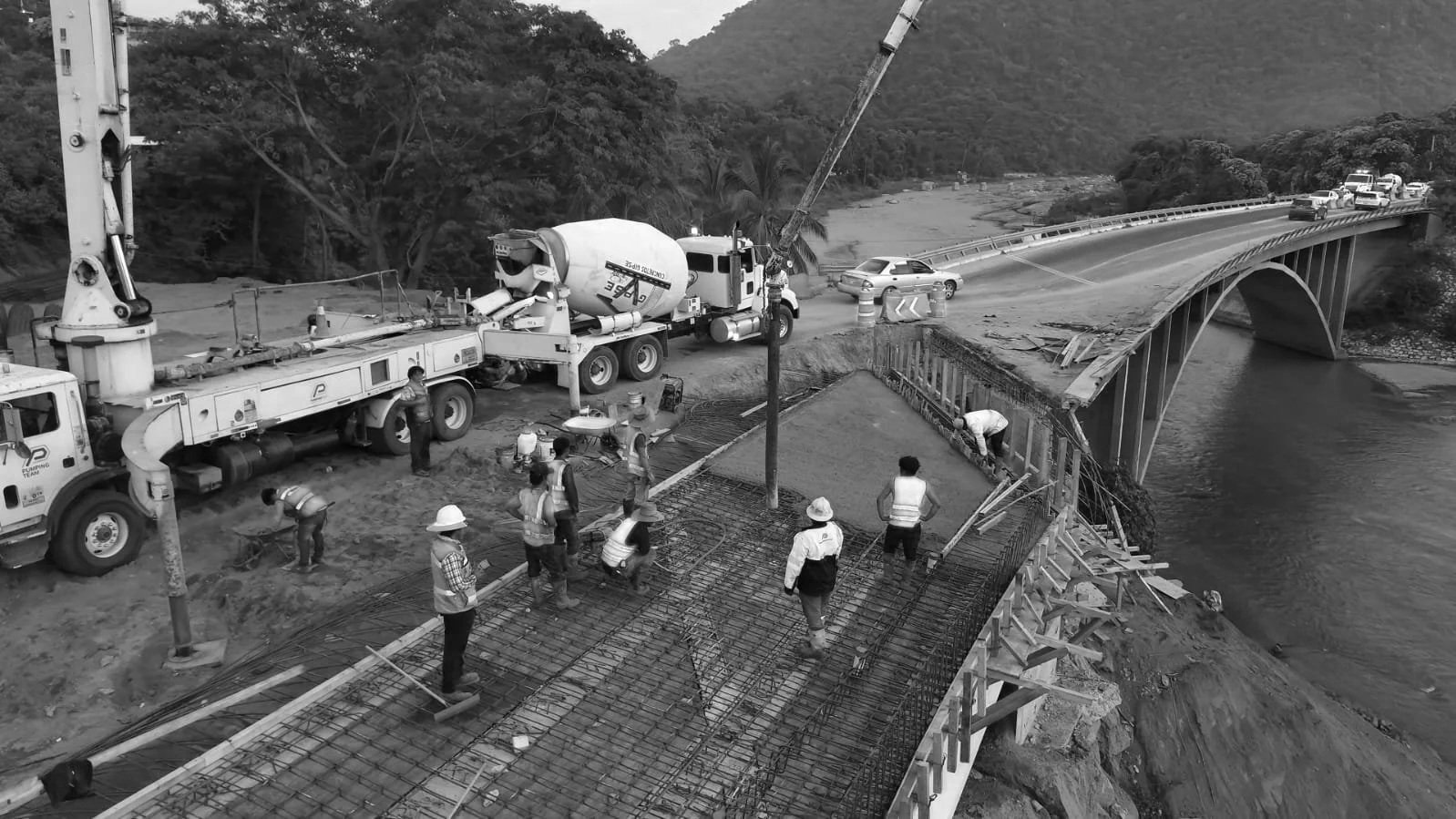 Construcción de un puente sobre un río en una zona montañosa. Trabajadores en la estructura del puente, maquinaria pesada y un camión mezclador de cemento.