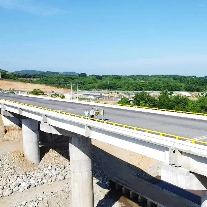 Una vista de un puente en construcción o reciente, con tres personas de pie en la superficie del puente, rodeado de un paisaje verde y montañoso bajo un cielo despejado.