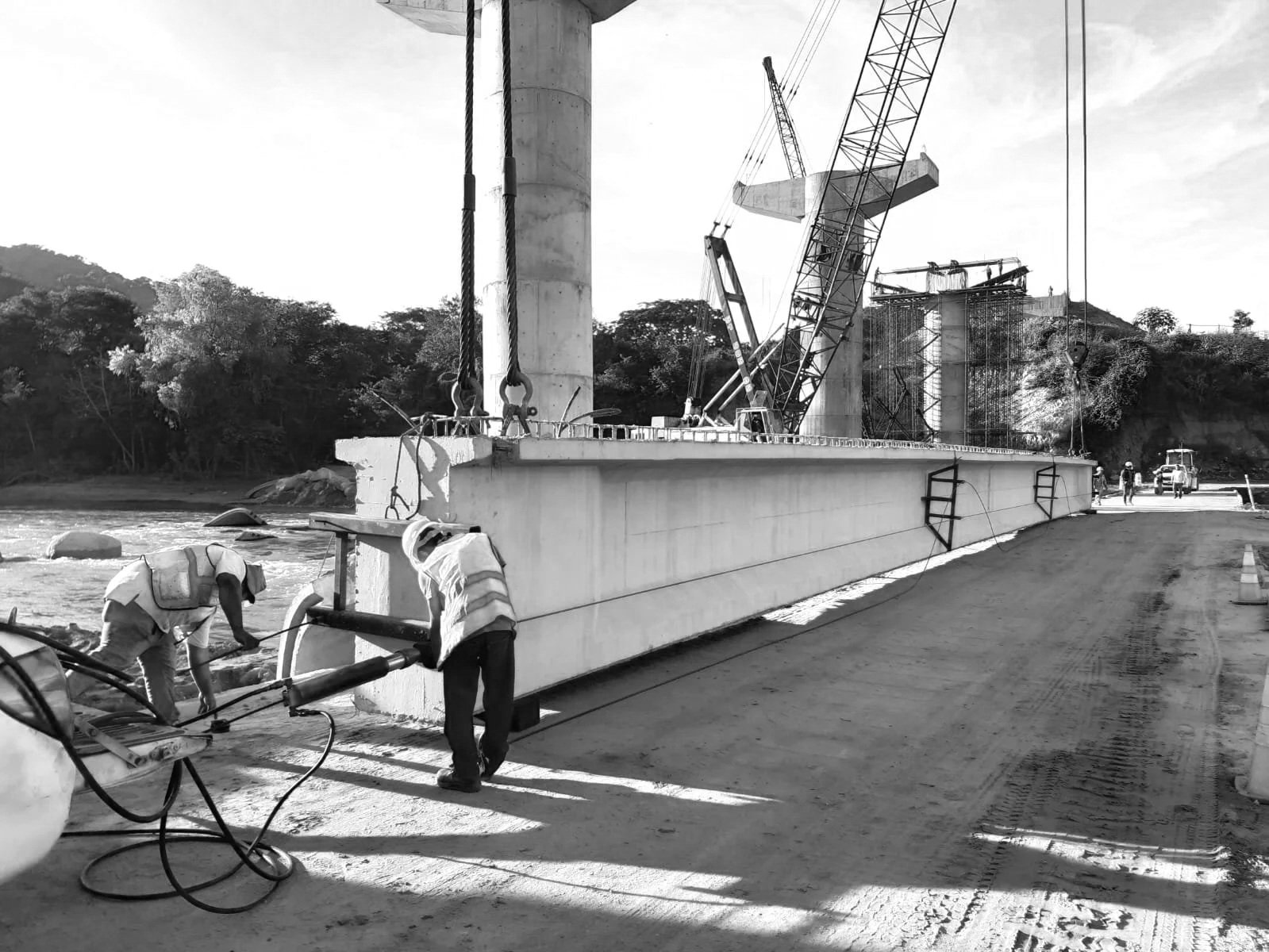 Trabajadores en un sitio de construcción levantando una estructura grande con grúas en un río, con árboles al fondo y maquinaria en el fondo.