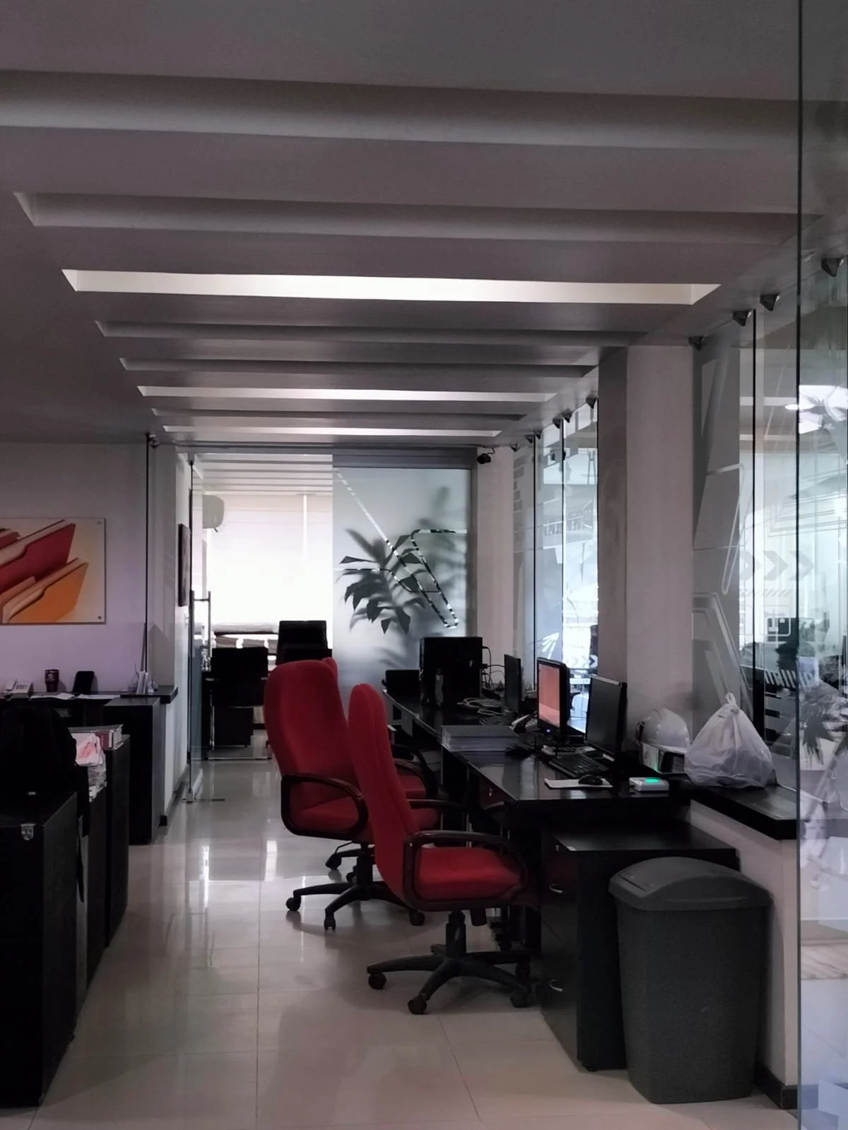Oficina con escritorios, sillas rojas y negras, computadoras, computadora portátil, plantas, y ventanas de vidrio con vista a la calle.