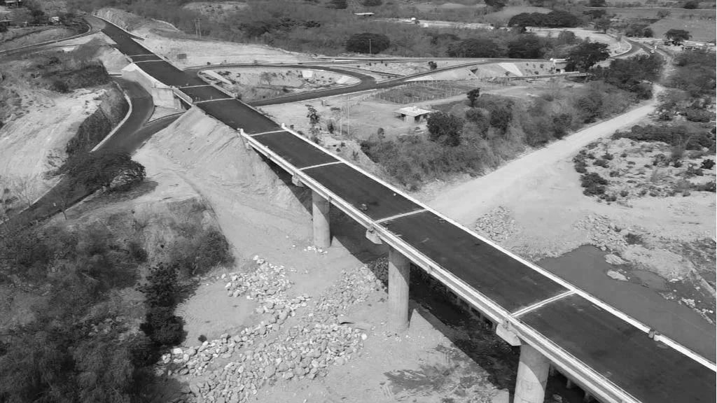 Puente en construcción sobre río, rodeado de tierra y vegetación, vista desde arriba en un área rural.