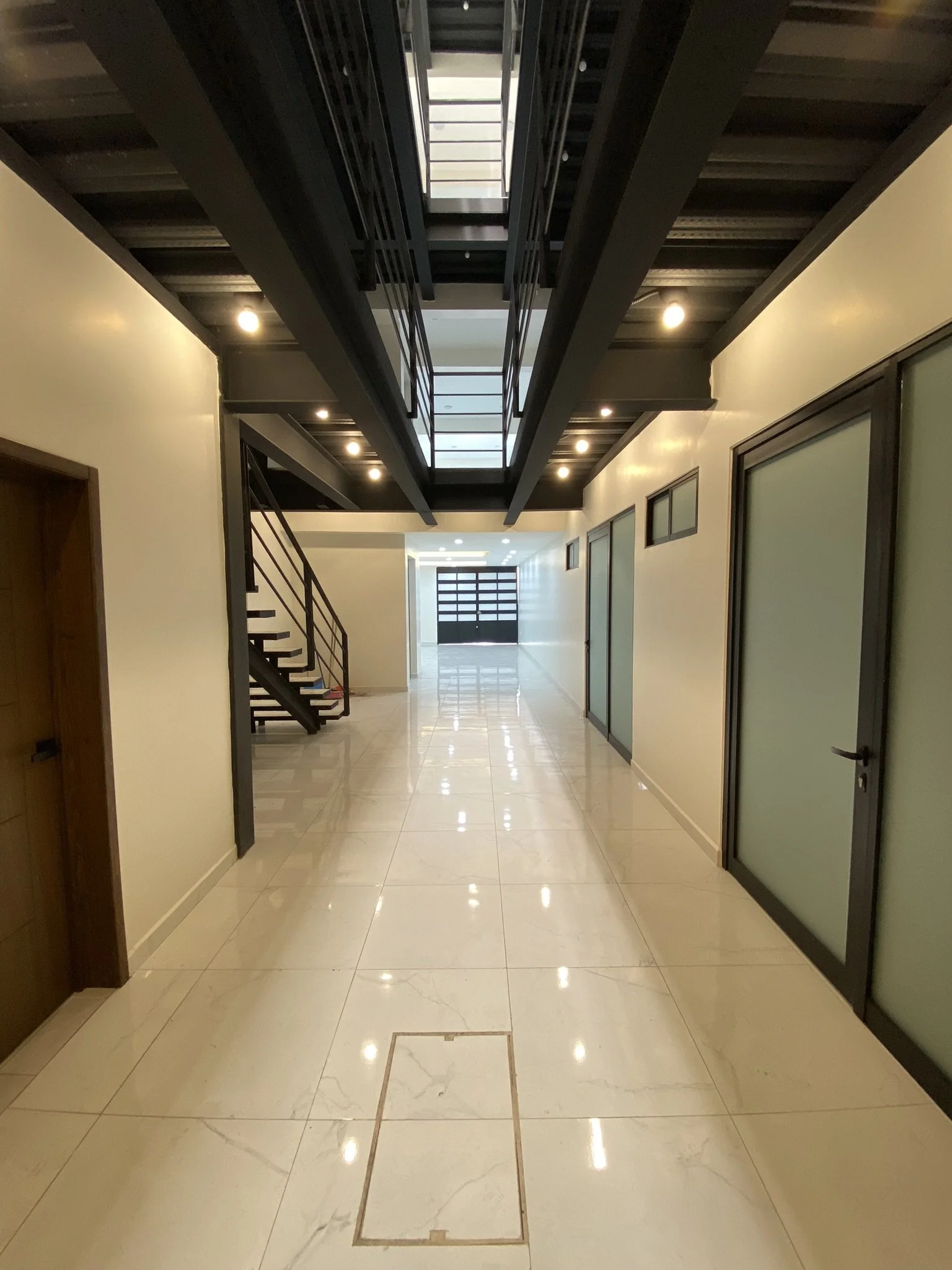 Pasillo en un edificio con paredes claras, puertas con cristales esmerilados a ambos lados, piso de baldosas blancas con vetas grises, escaleras de madera y metal a la izquierda y ventanas en la pared derecha, luminarias en el techo.