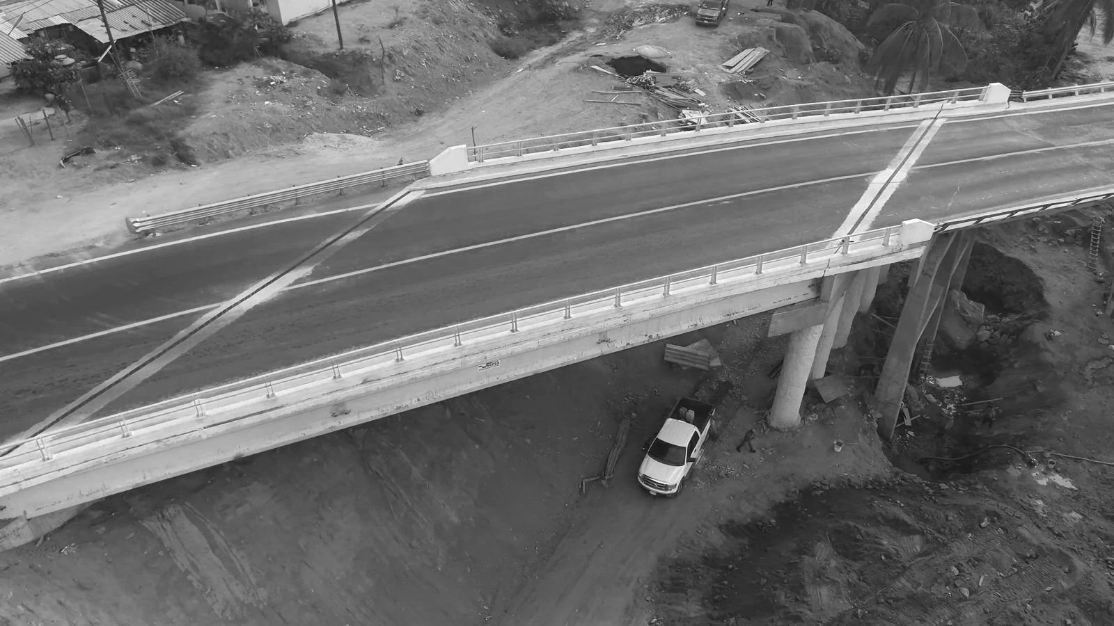 Imagen en blanco y negro de un puente en construcción, con estructura de concreto y barandas, sobre un terreno de tierra, con maquinaria y vehículos en el área de construcción.