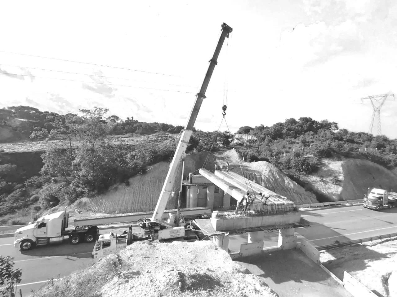 Trabajadores ensamblando una estructura de puente con una grúa en una carretera rodeada de naturaleza y colinas.