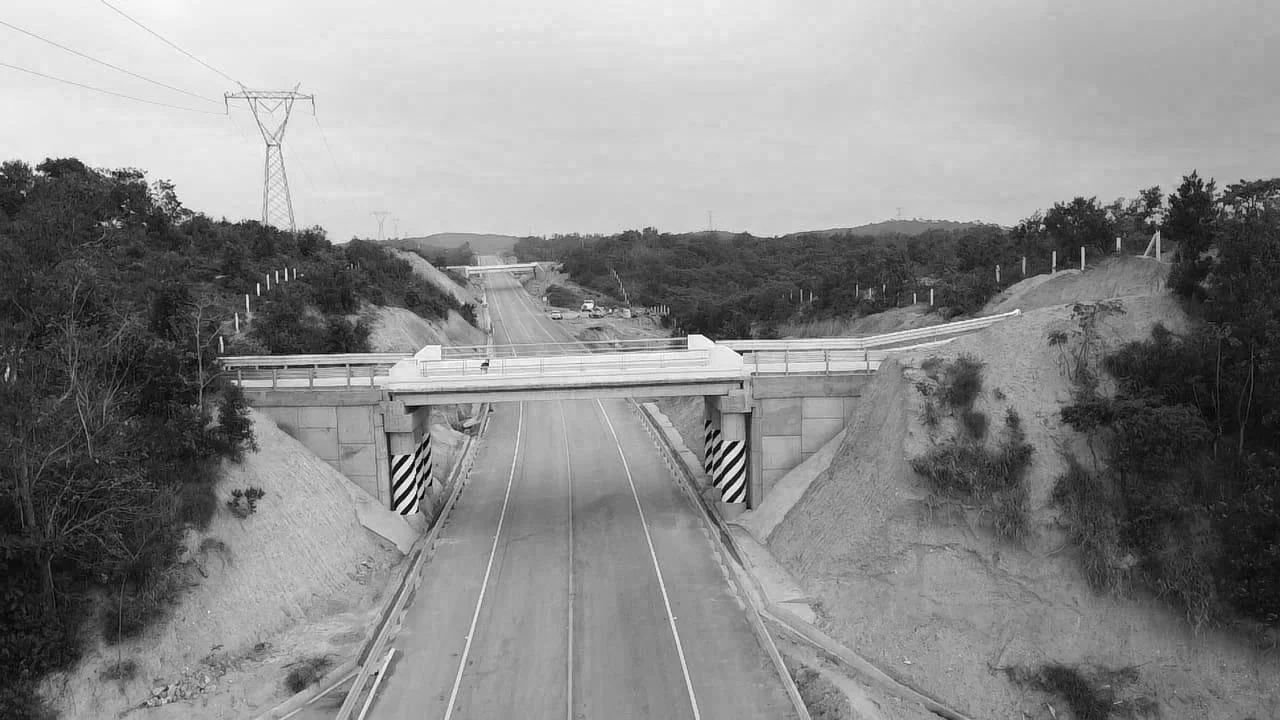 Puente vial en una vía de dos carriles, rodeado de terreno montañoso con vegetación y líneas eléctricas en el fondo.