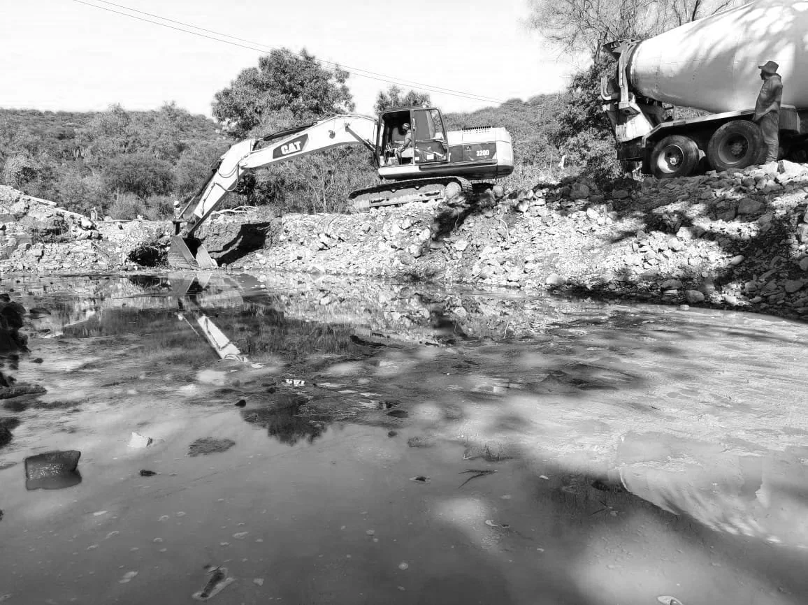 Maquinaria de construcción operando junto a un río con aguas tranquilas, en un área de tierra y rocas, en un ambiente rural con árboles y cables sobre el cielo.