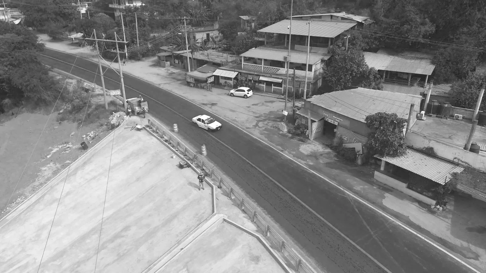 Vista aérea en blanco y negro de una calle con dos autos circulando, flanqueada por casas y árboles, con postes de electricidad y un camión de construcción en una esquina.