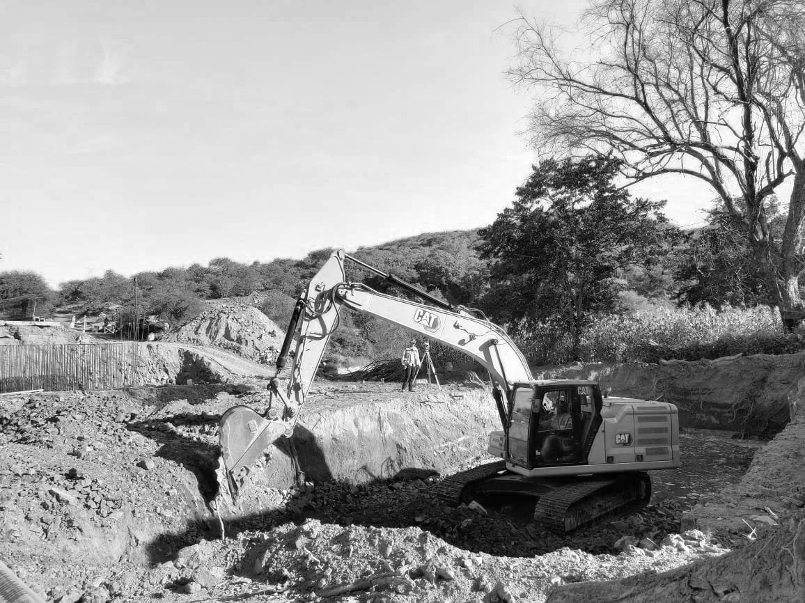 Carriola excavadora trabajando en una excavación en un sitio de construcción con árboles en el fondo y dos personas observando.