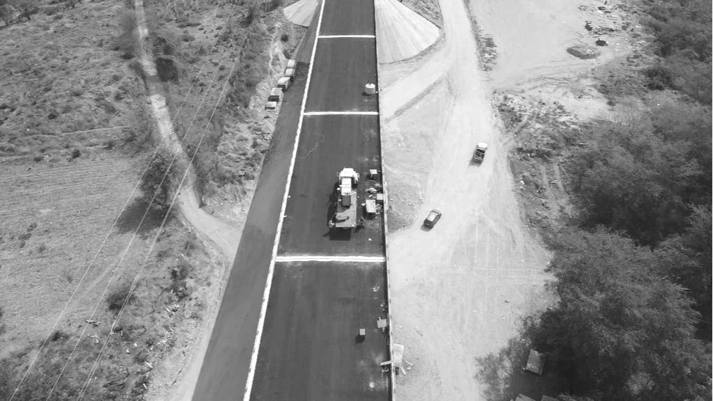 Vista aérea en blanco y negro de una calle en construcción, con vehículos y maquinaria trabajando en una mitad y terreno sin pavimentar en la otra mitad.