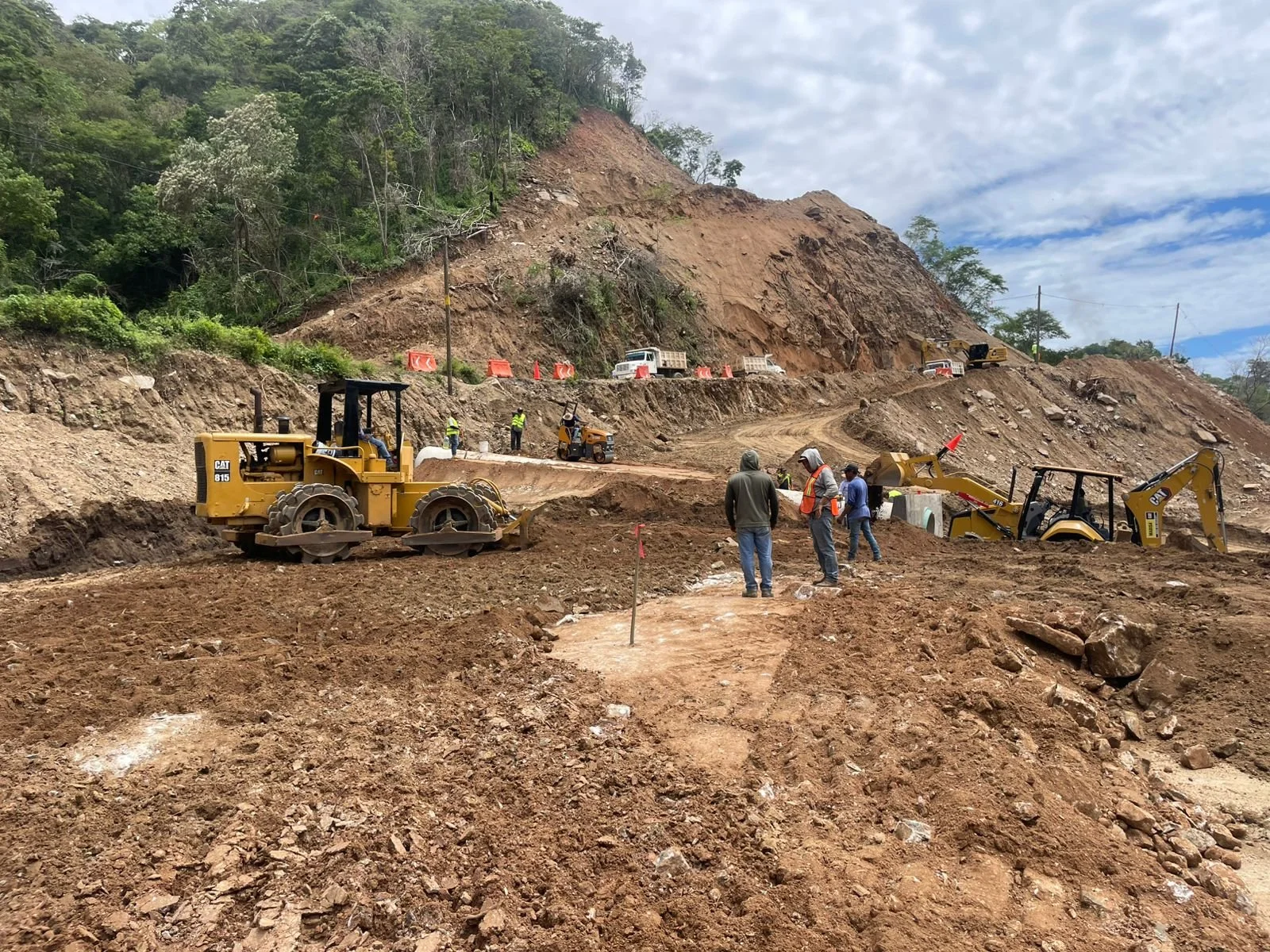Obra de construcción en una carretera de montaña. Maquinaria pesada como excavadoras y rodillos compactadores, trabajadores con cascos y chalecos de seguridad, y una ladera en proceso de estabilización o ampliación.
