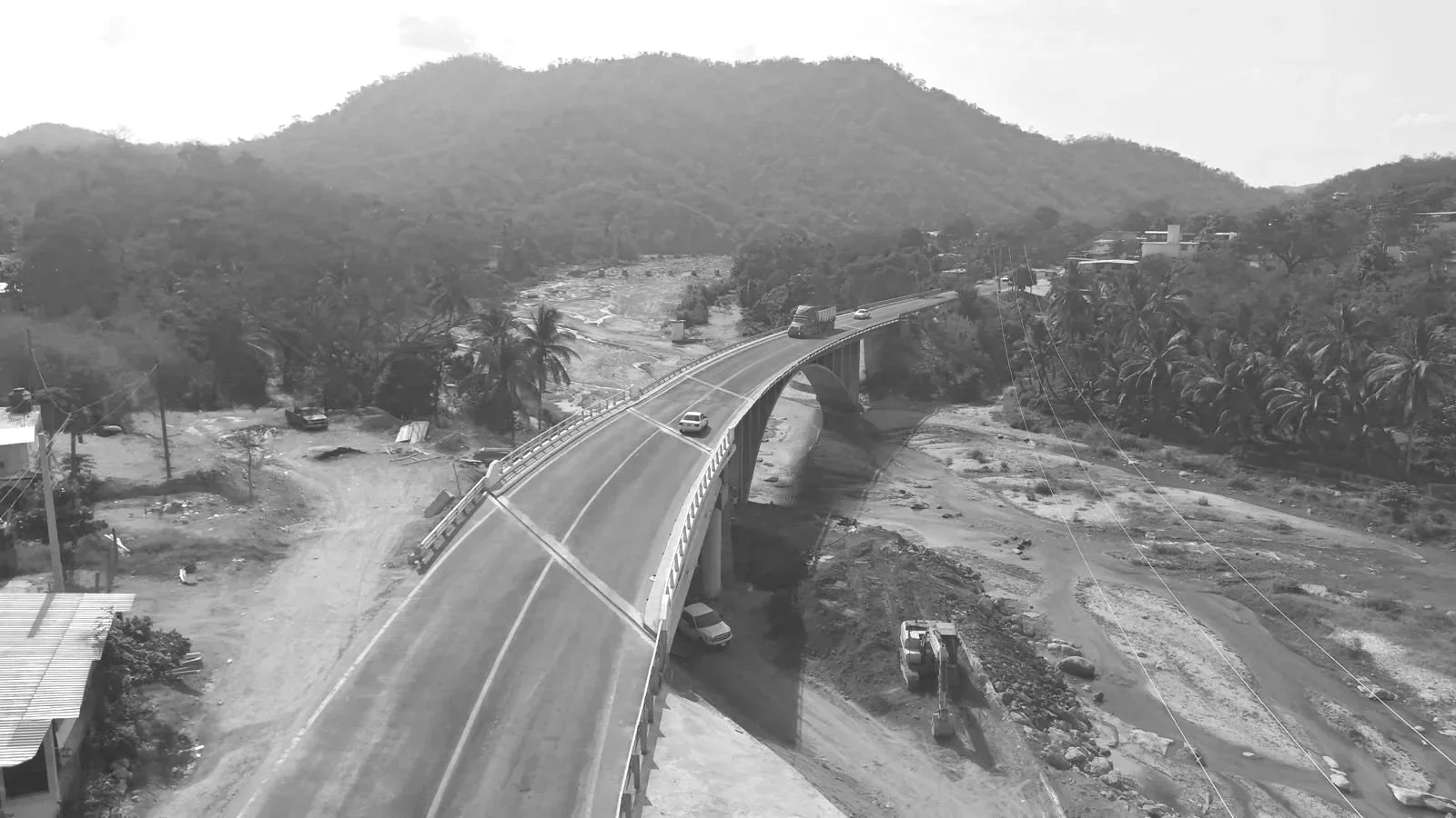 Puente en construcción sobre río en un área montañosa, con maquinaria de construcción y pocos vehículos en la carretera.