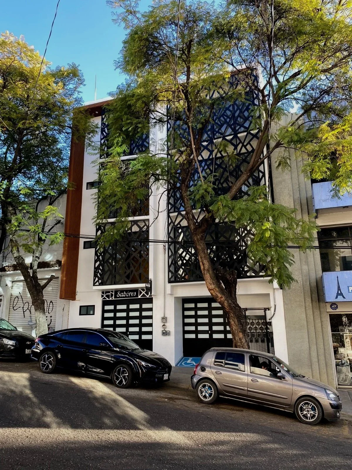 Edificio moderno con fachada decorativa de paneles negros en forma de cuadros y madera, con árboles en la calle y varios autos estacionados.