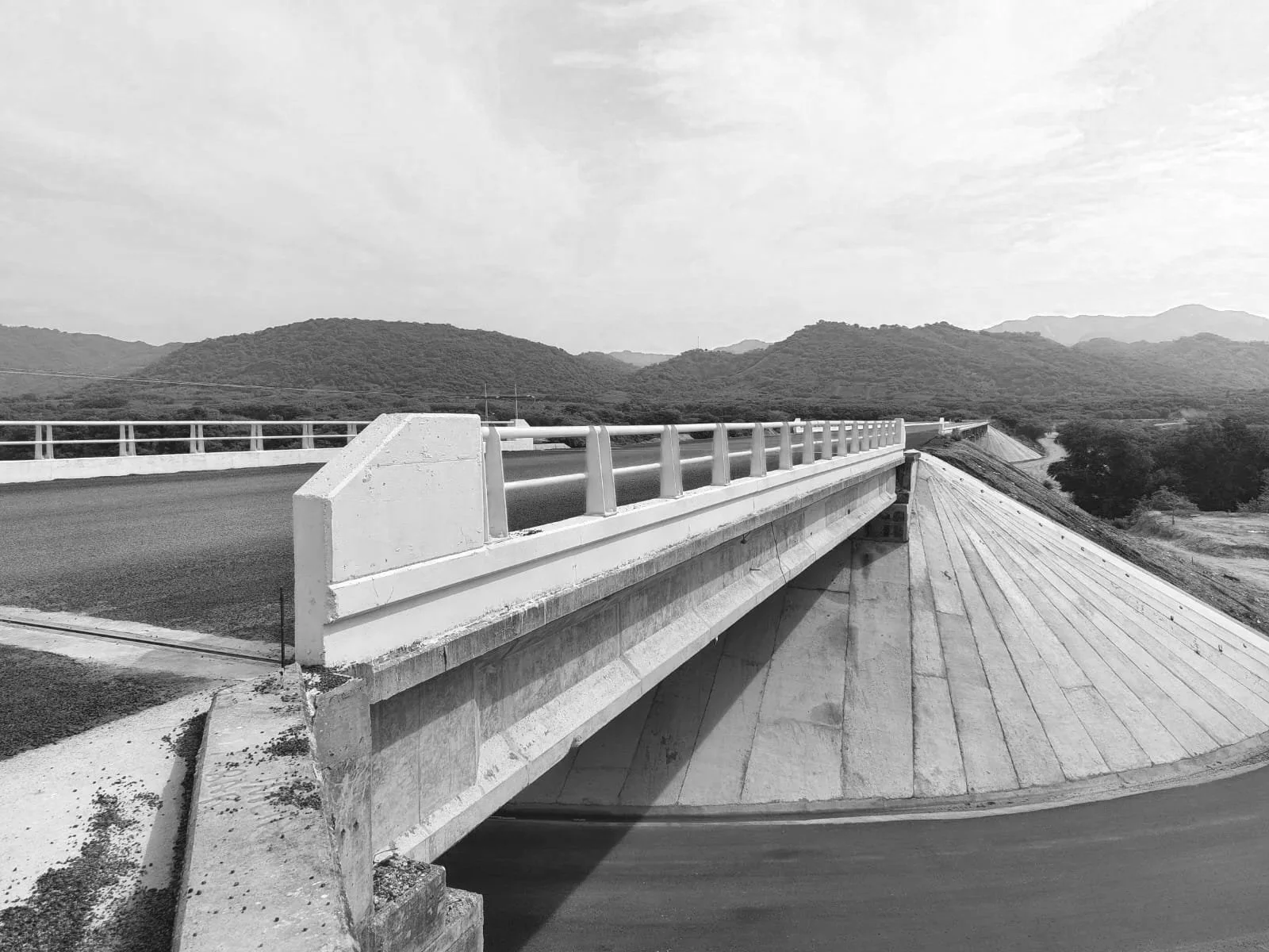Puente de concreto y metal sobre una carretera en un paisaje de montañas y cielo nublado, en escala de grises.
